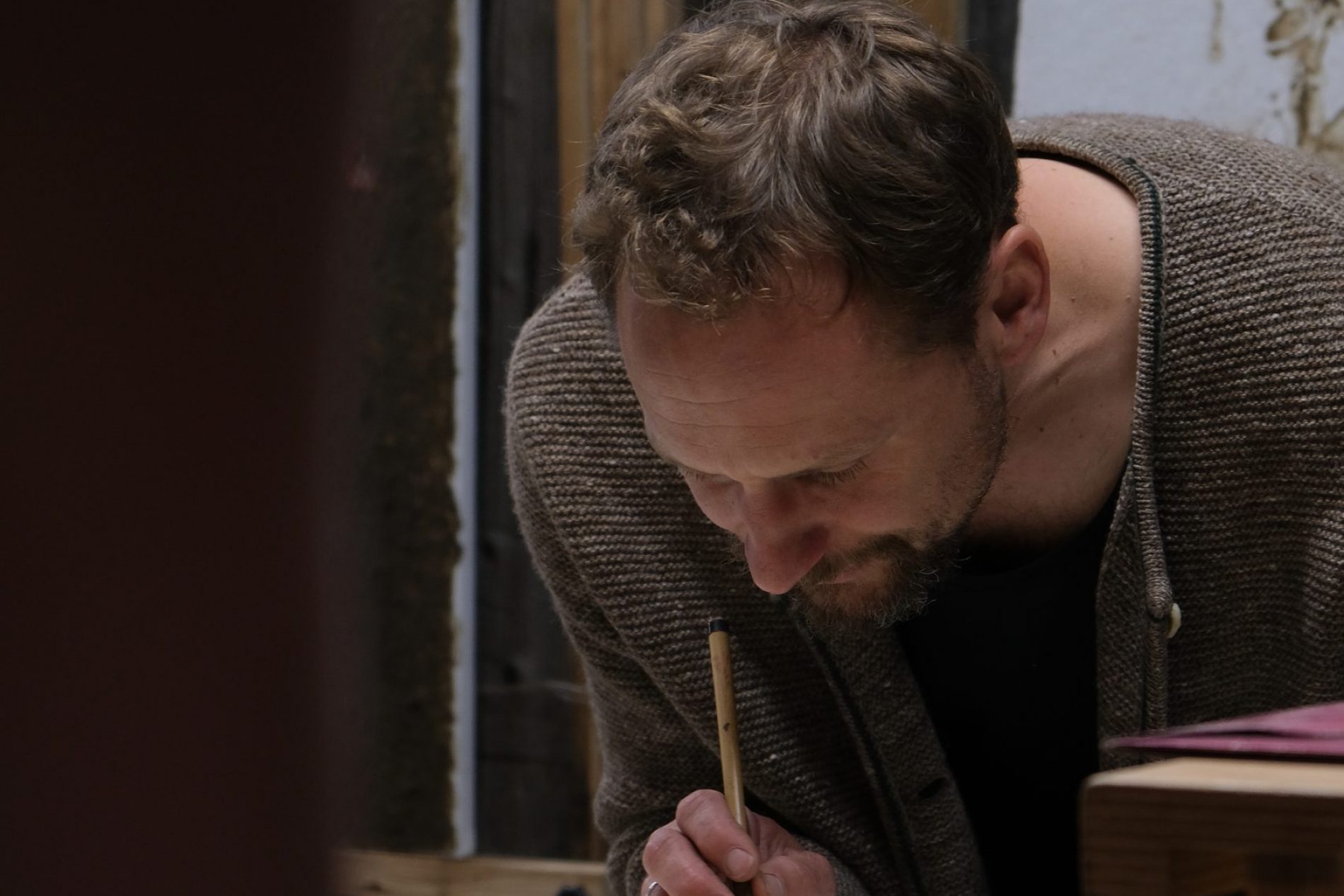 Mann in braunem Wollcardigan malt mit Pinsel auf einer Skizze; Atelier, Spezlcamp-Erfolgsgeschichte.