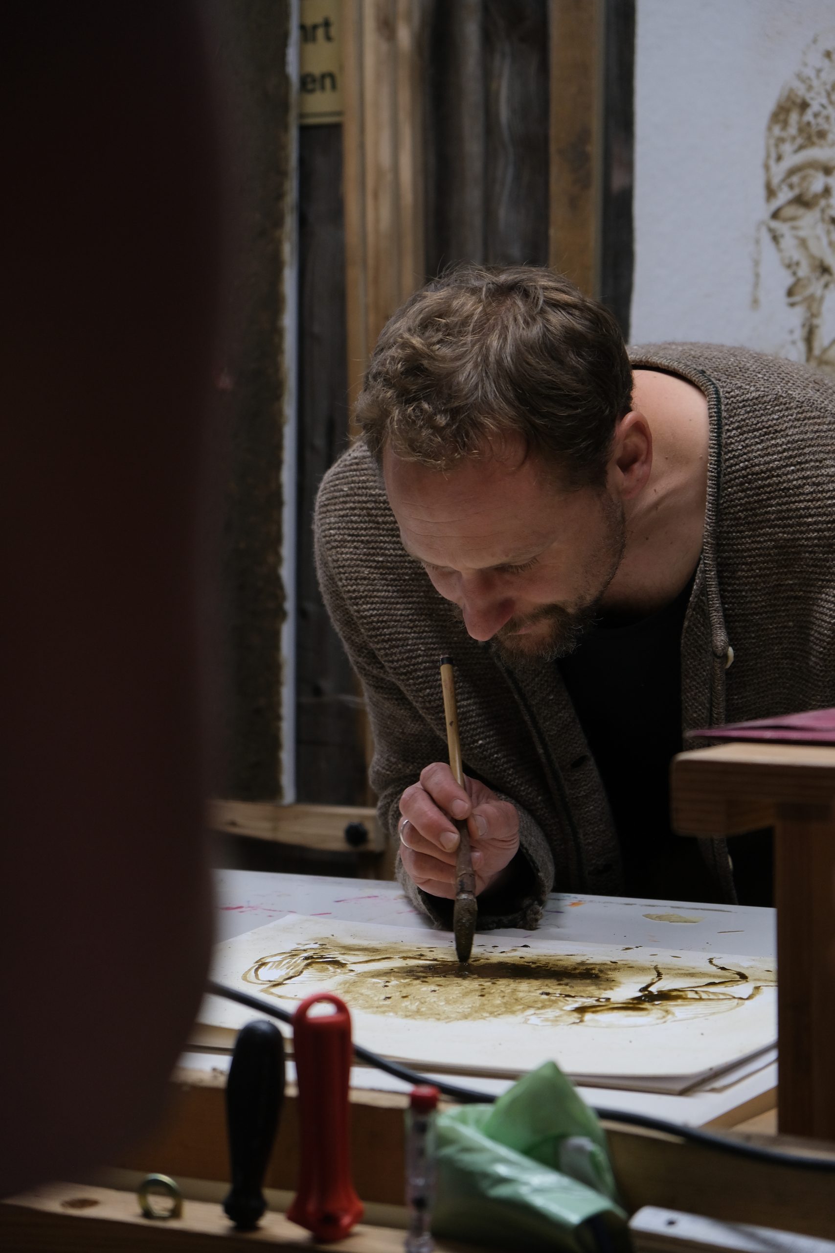 Mann in braunem Wollcardigan malt mit Pinsel auf einer Skizze; Atelier, Spezlcamp-Erfolgsgeschichte.