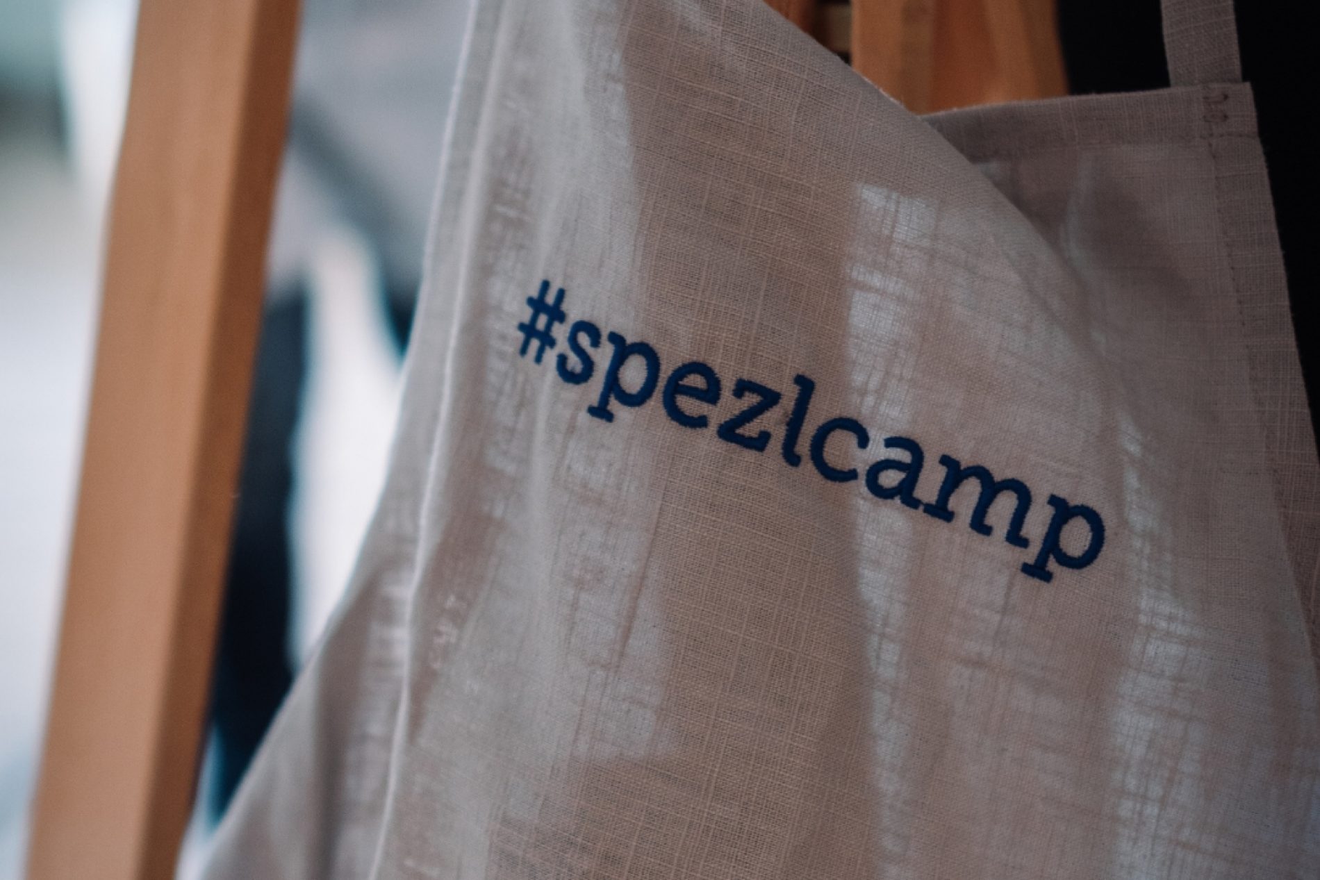 spezlcamp Stofftasche hängt an Holzrahmen; Text im Vordergrund, unscharfer Hintergrund.
