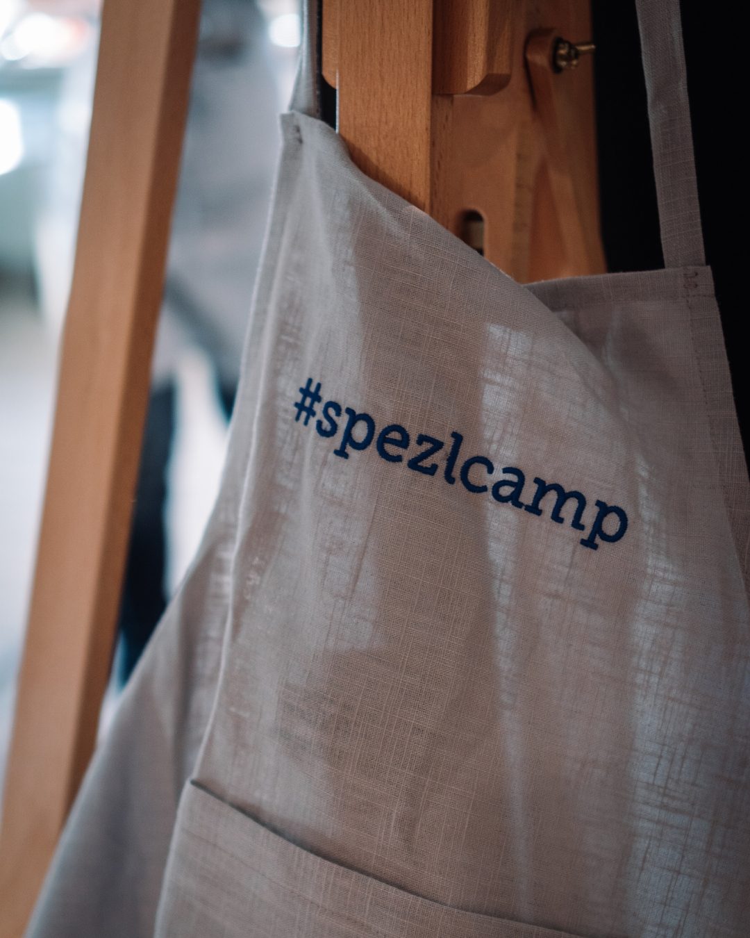 spezlcamp Stofftasche h&auml;ngt an Holzrahmen; Text im Vordergrund, unscharfer Hintergrund.