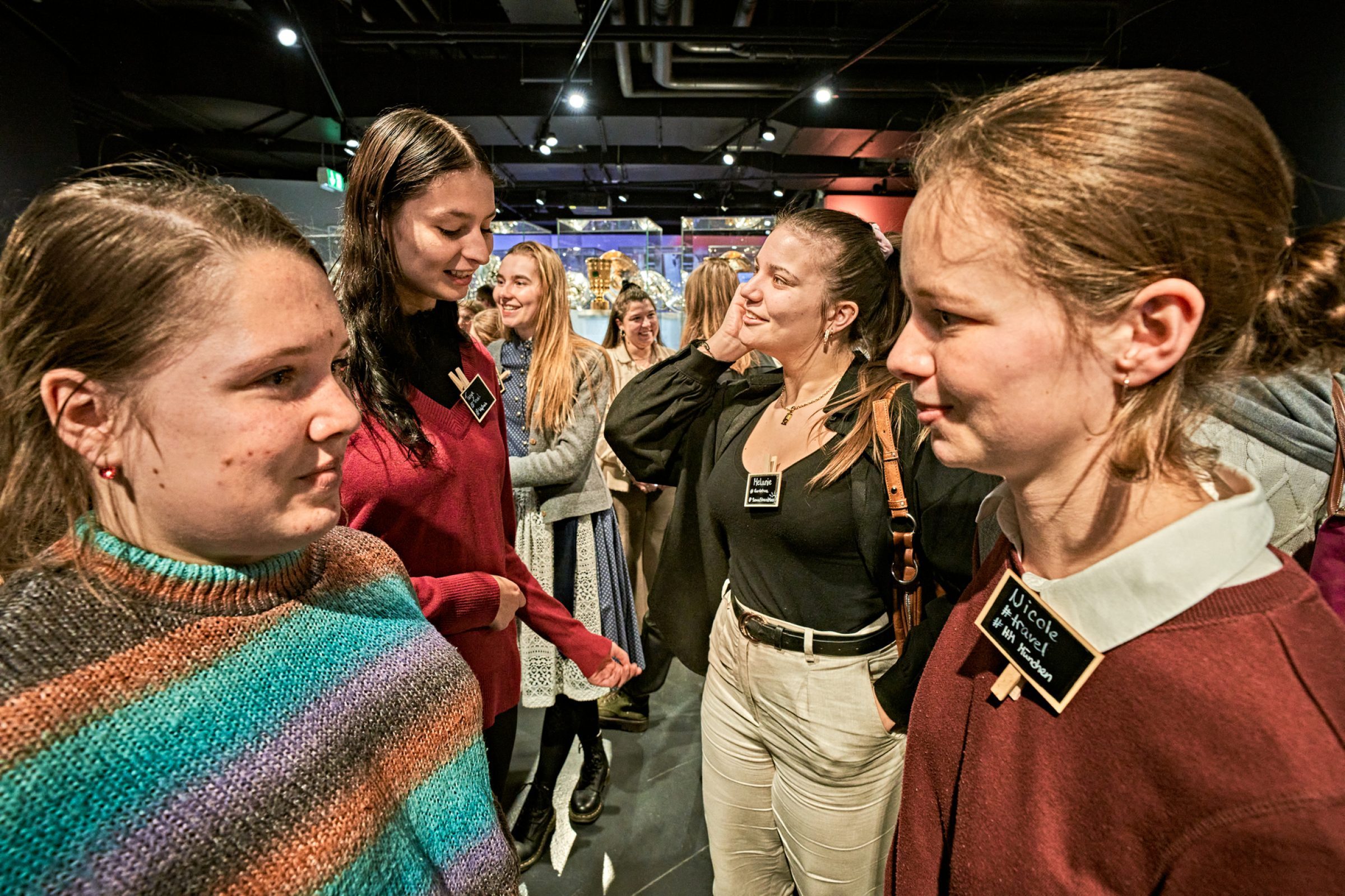 Gruppe junger Menschen mit Namensschildern im Gespr&auml;ch auf einer Messefl&auml;che; Ausstellungsst&auml;nde im Hintergrund, Tourism Next
