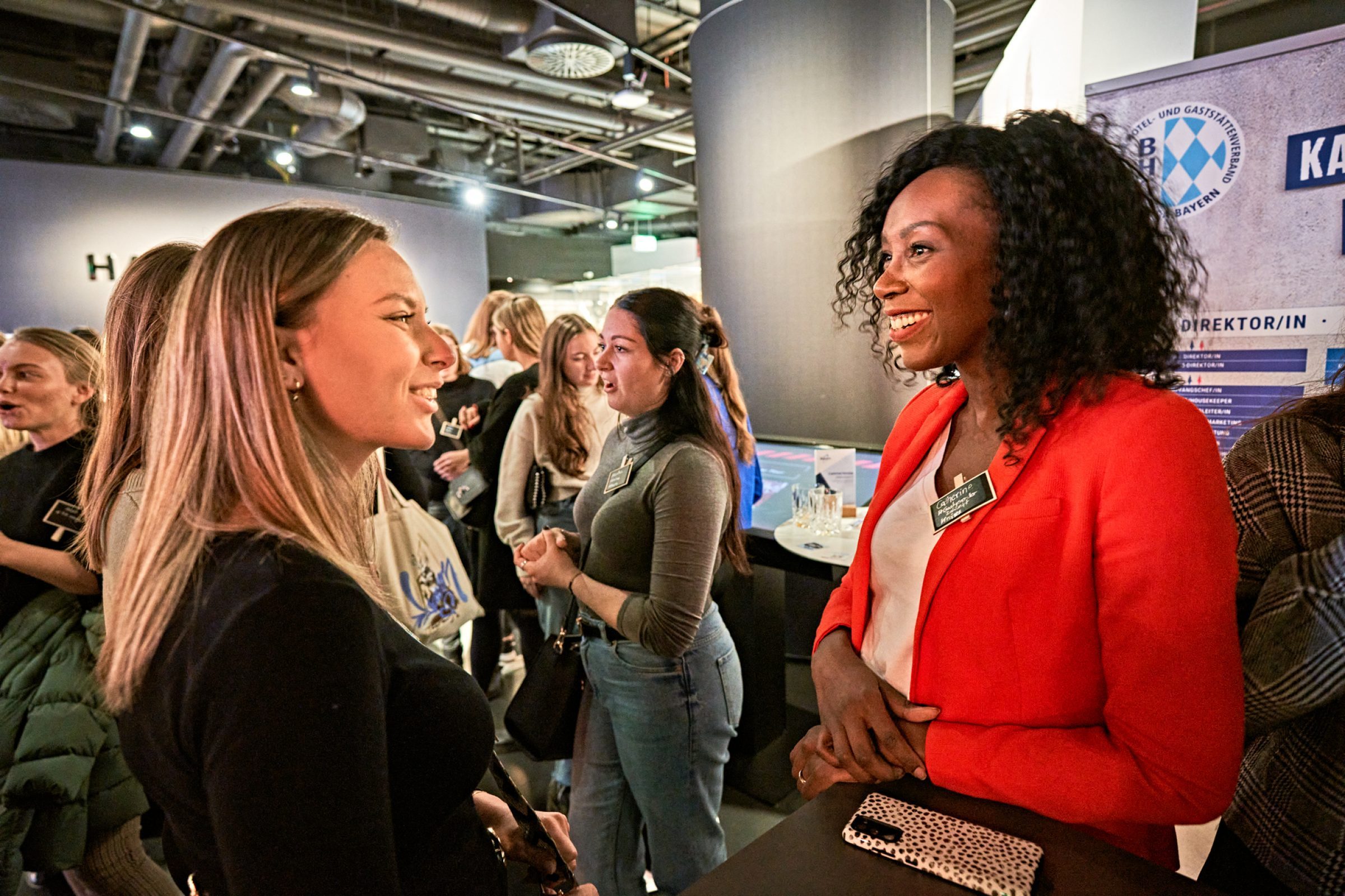 Networking auf Tourism Next in der Allianz Arena: zwei Frauen im Gespr&auml;ch am Messestand, weitere Teilnehmer im Hintergrund.