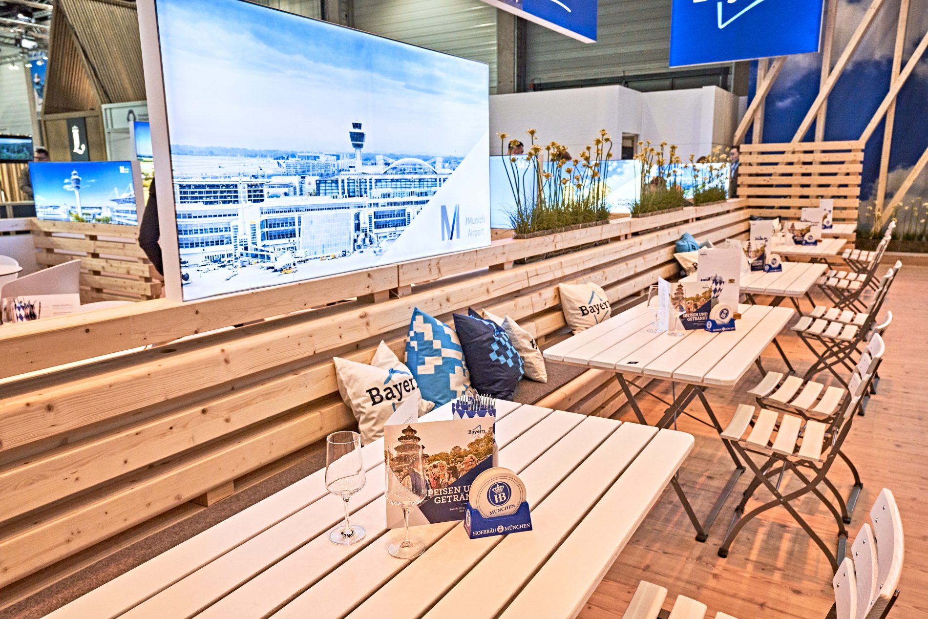 Bayern-Stand ITB 2025: lange Holztische mit Bänken, Bildschirmwand, Pflanzenspaliere, Bayern-Kissen und Info-Material.