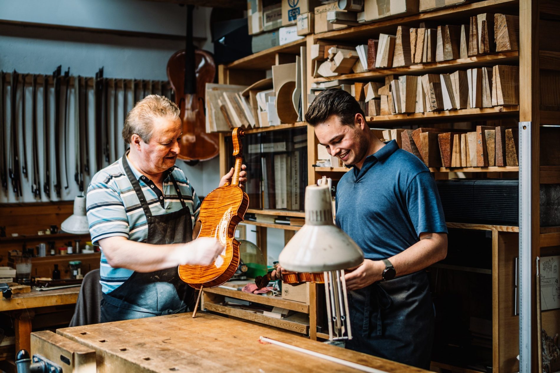 Zwei Männer in einer Werkstatt bearbeiten eine Violine; im Hintergrund Holzkisten, Holzleisten und Werkzeugregale.