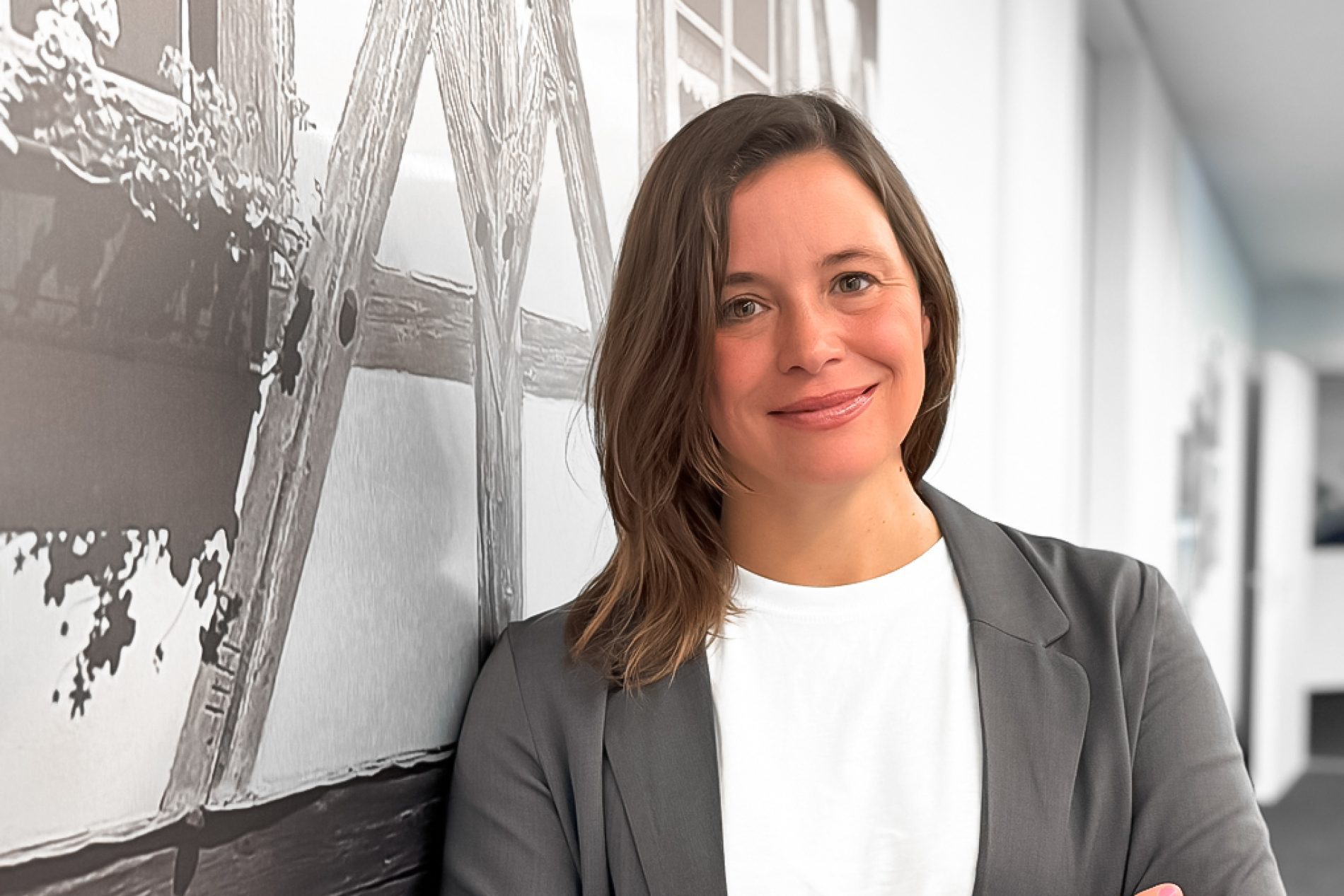 Isabella Hansch Isabella Hansch lächelt, grauer Blazer, weißes T-Shirt; steht an Wand mit Holzbalken-Fotodruck im Büro.