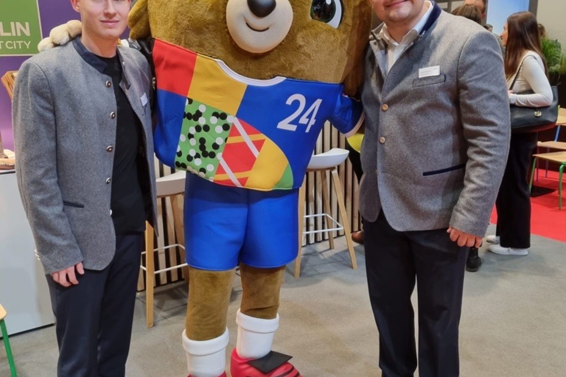 Zwei Junge Menschen stehen neben einem großen Stoff-Maskottchen der Europameisterschaft 2024.