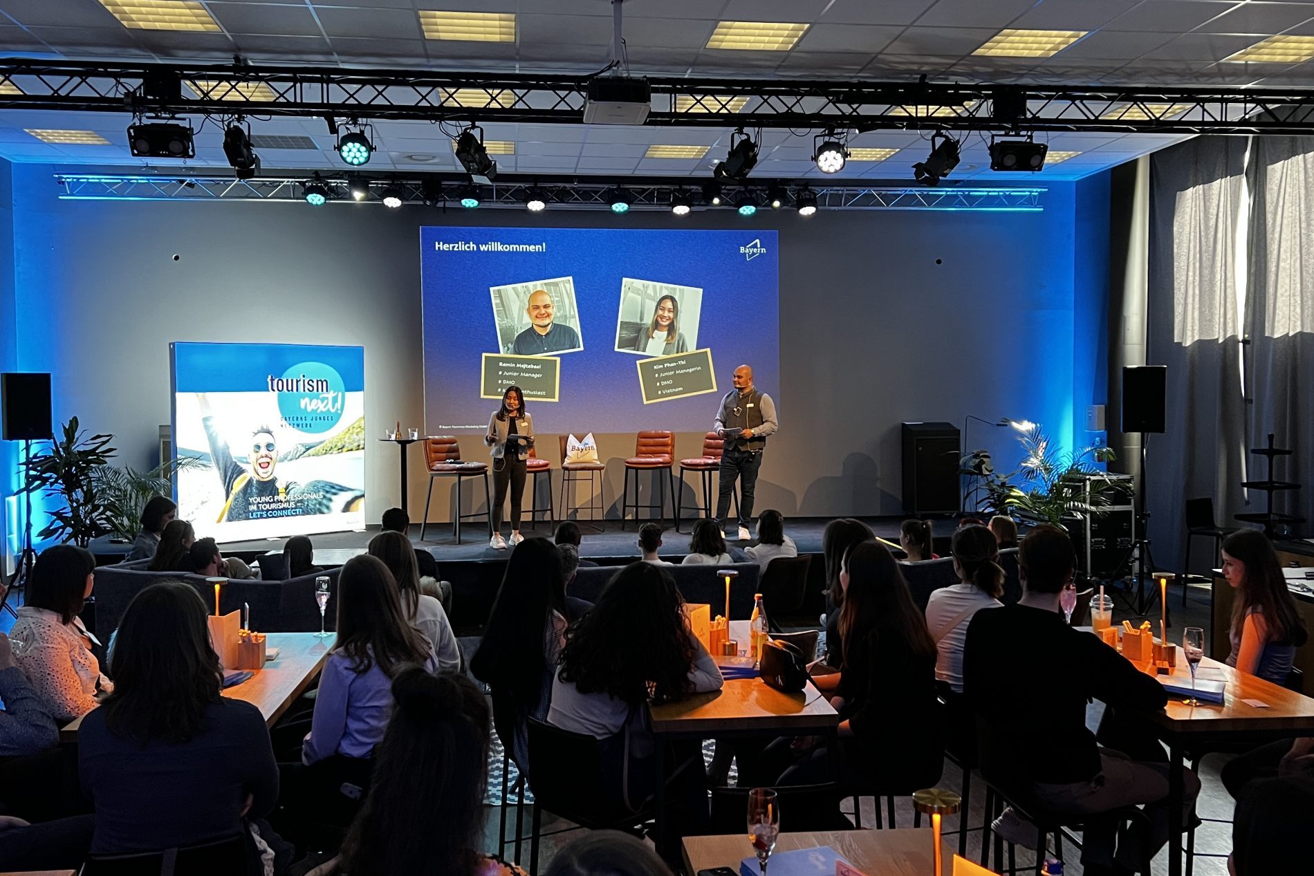 B&uuml;hne mit zwei Moderatoren bei Tourism Next in Regensburg, gro&szlig;er Bildschirm mit Portraits, Publikum an Tischen