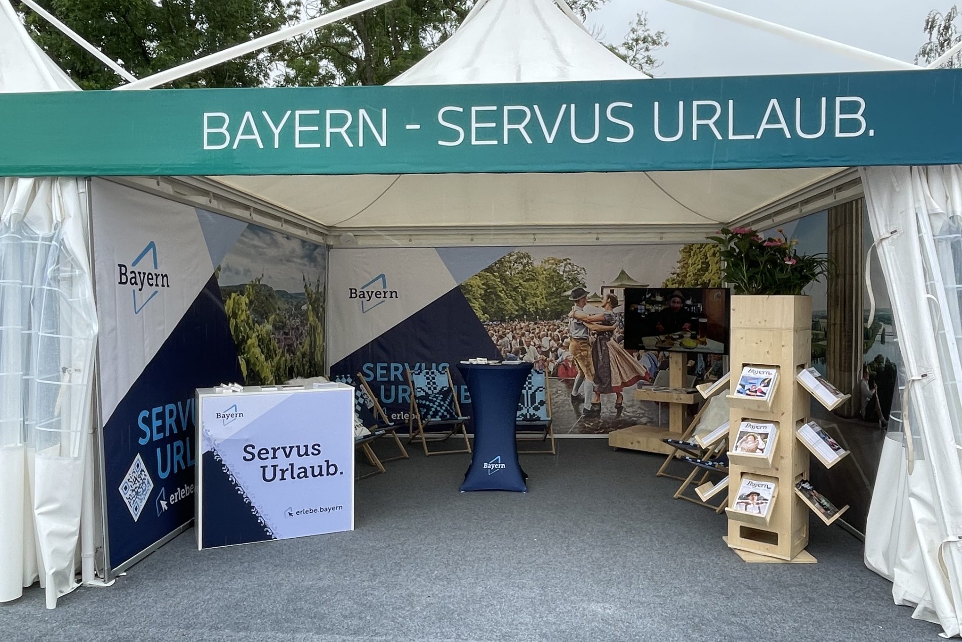 Zeltstand Bayern mit Banner BAYERN - SERVUS URLAUB. Infotisch, Brosch&uuml;renregal, Sitzbereich, TV-Bildschirm und Pflanzen.
