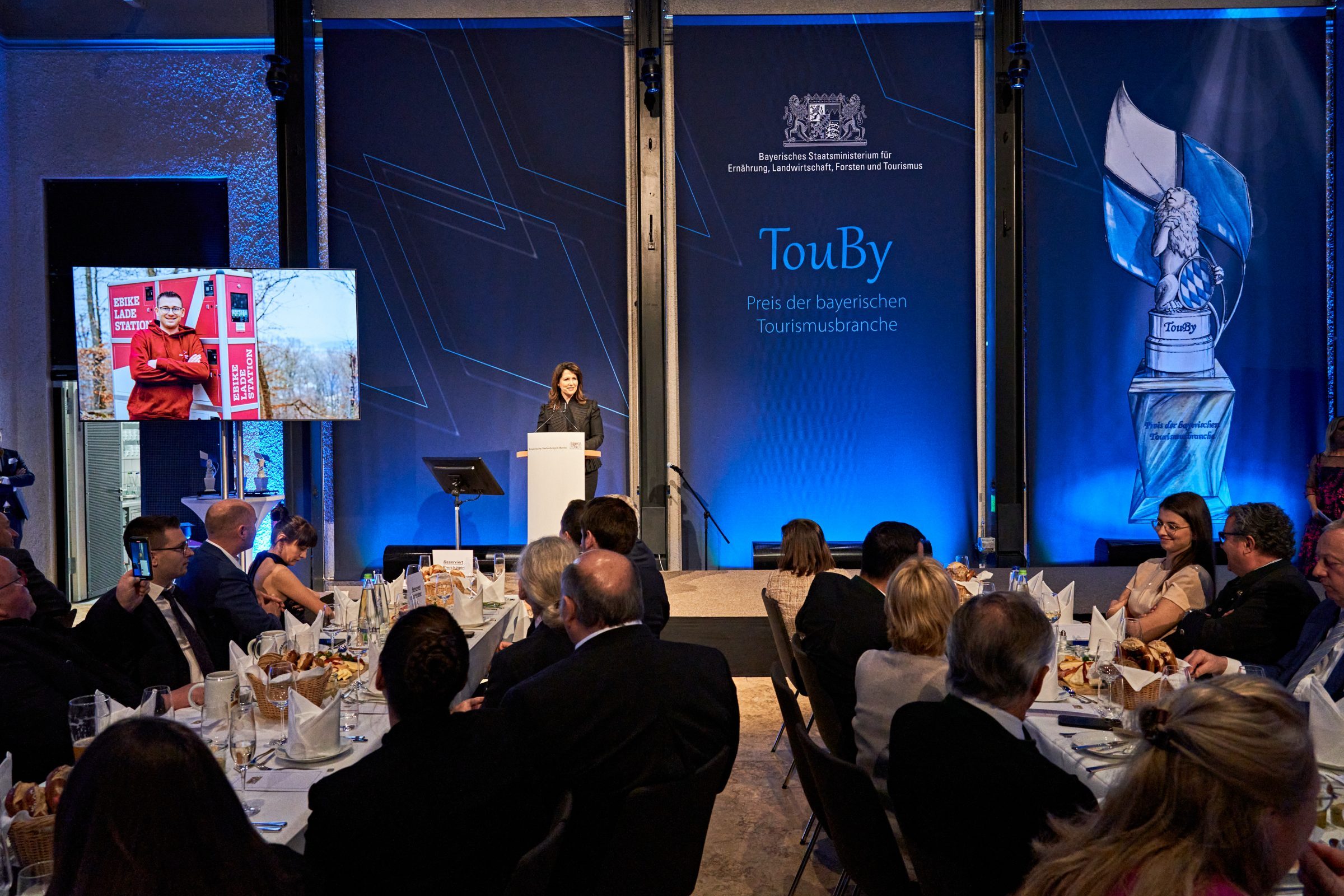 Blick in einen Veranstaltungsraum, in dem Menschen im Vordergrund an Tischen sitzen. Auf der B&uuml;hne steht eine Frau an einem Rednerpult. Der Hintergrund ist blau mit der Aufschrift "TouBy".