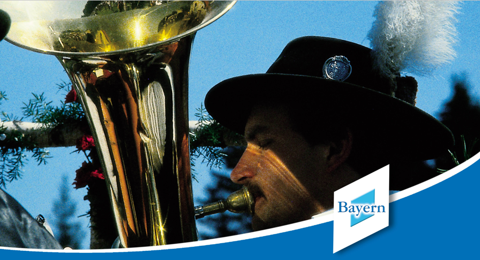 0_Titelbild_Option1_(c)BayTM Eine Mann spielt ein Blasinstrument, er trägt einen traditionellen Hut mit einer Feder daran. Im Vordergrund ist ein blauer Balken und unser altes Logo mit dem Schriftzug "Bayern" zu sehen.