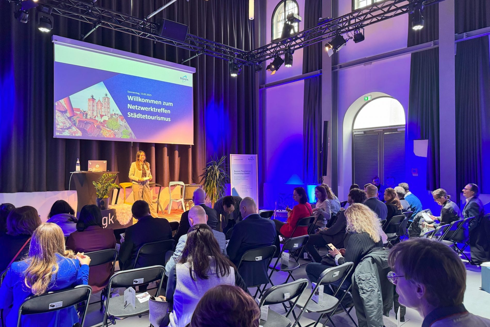 Moderatorin auf Bühne beim Netzwerktreffen Städtetourismus Ingolstadt; Projektion zeigt Willkommenstext, Publikum sitzt.