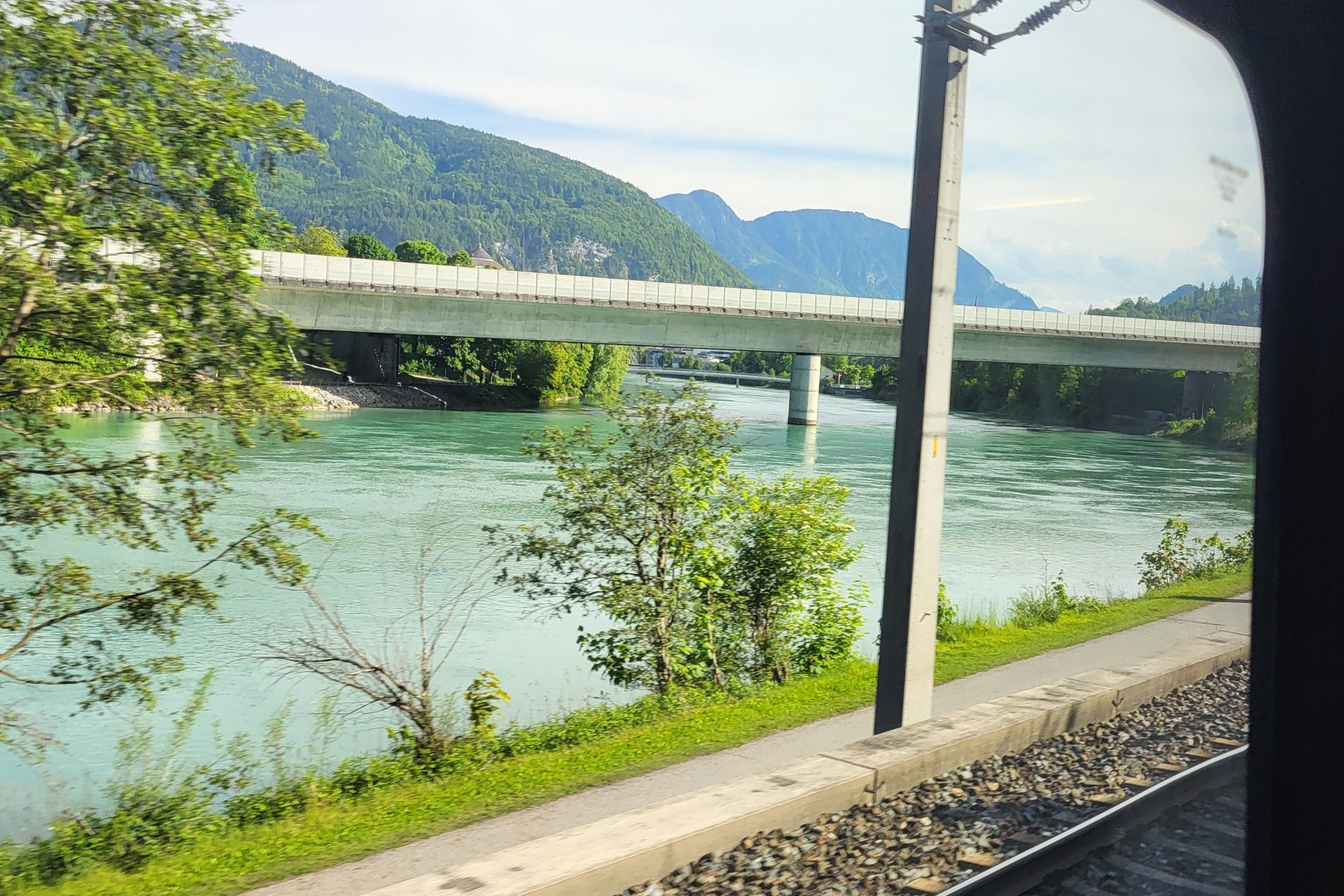 Blick aus dem Zugfenster auf Fluss, Brücke, Bahngleise, Uferweg, grüne Bäume und bergige Landschaft.