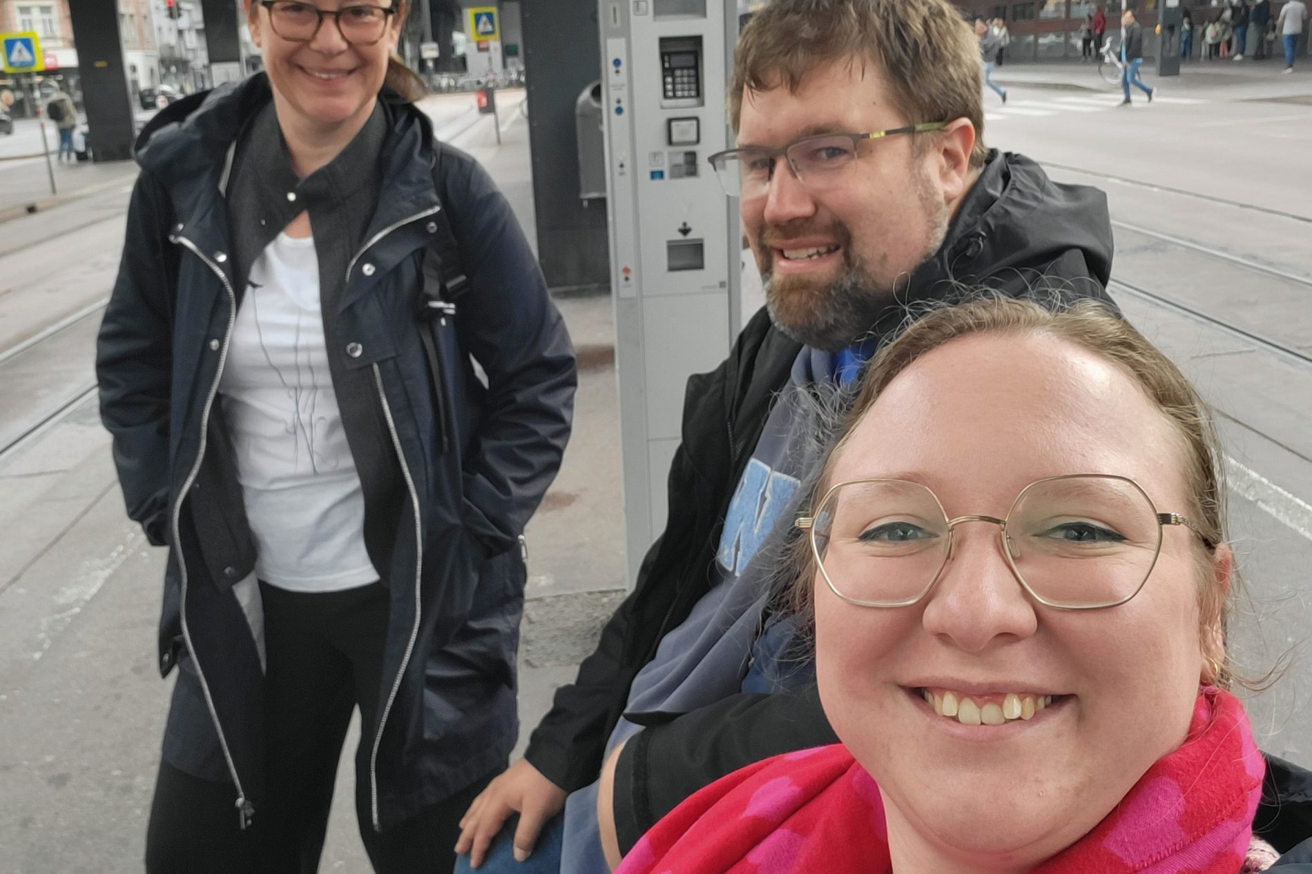 Drei Personen lachen am Busbahnhof neben Ticketautomat; urbane Umgebung, ODTA Treffen 2025.