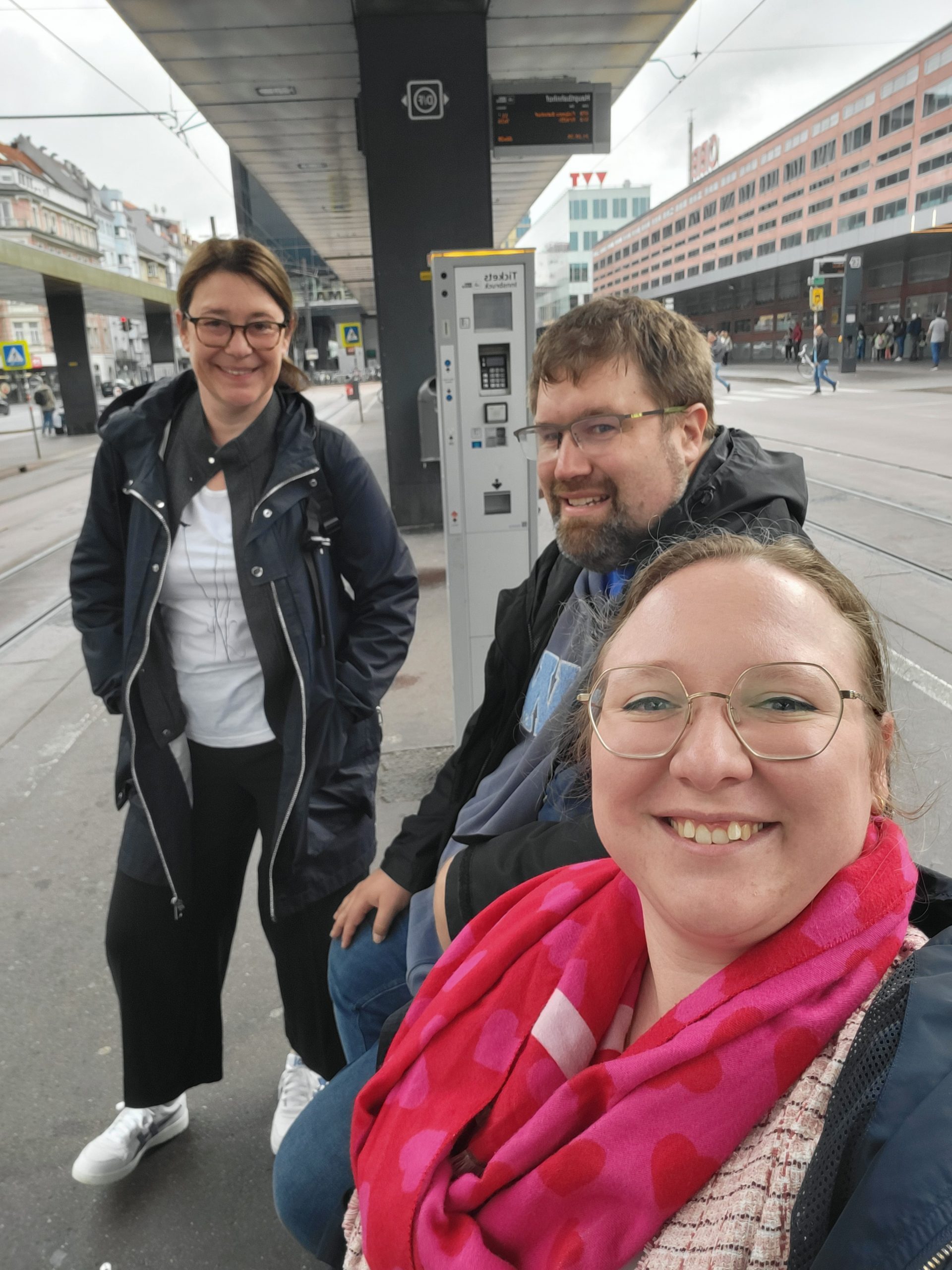 Drei Personen lachen am Busbahnhof neben Ticketautomat; urbane Umgebung, ODTA Treffen 2025.
