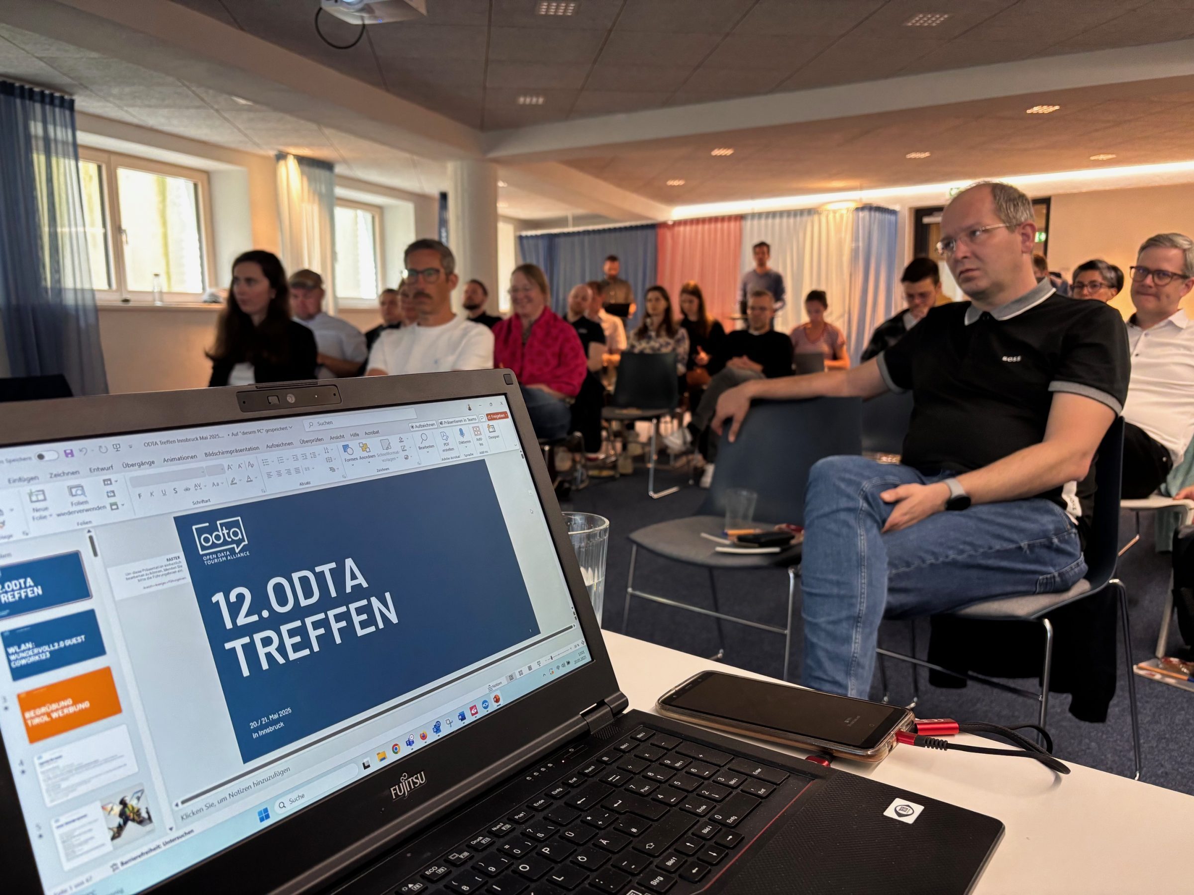 Laptop-Bildschirm zeigt Folie „12.ODTA TREFFEN“; im Hintergrund sitzen Teilnehmer bei ODTA-Treffen Innsbruck 2025.