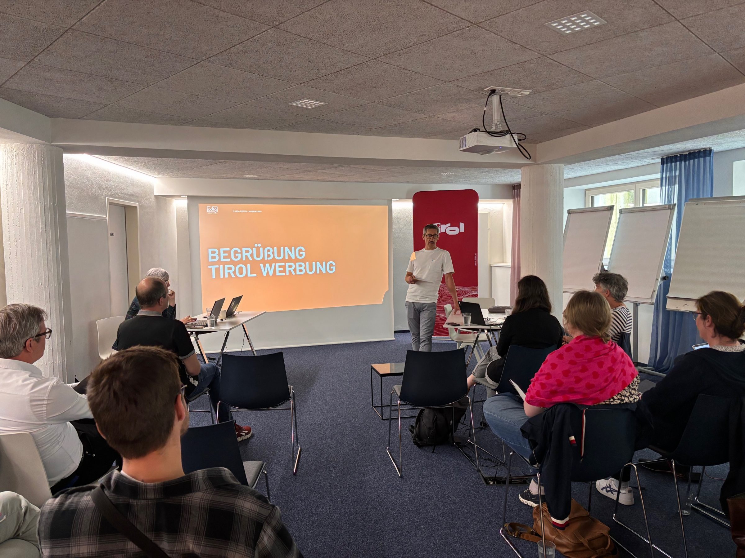 Vortragsraum beim ODTA Treffen Innsbruck 2025; Moderator vor Laptop, Bildschirm zeigt Begrüßung Tirol Werbung, Zuhörer sitzen.