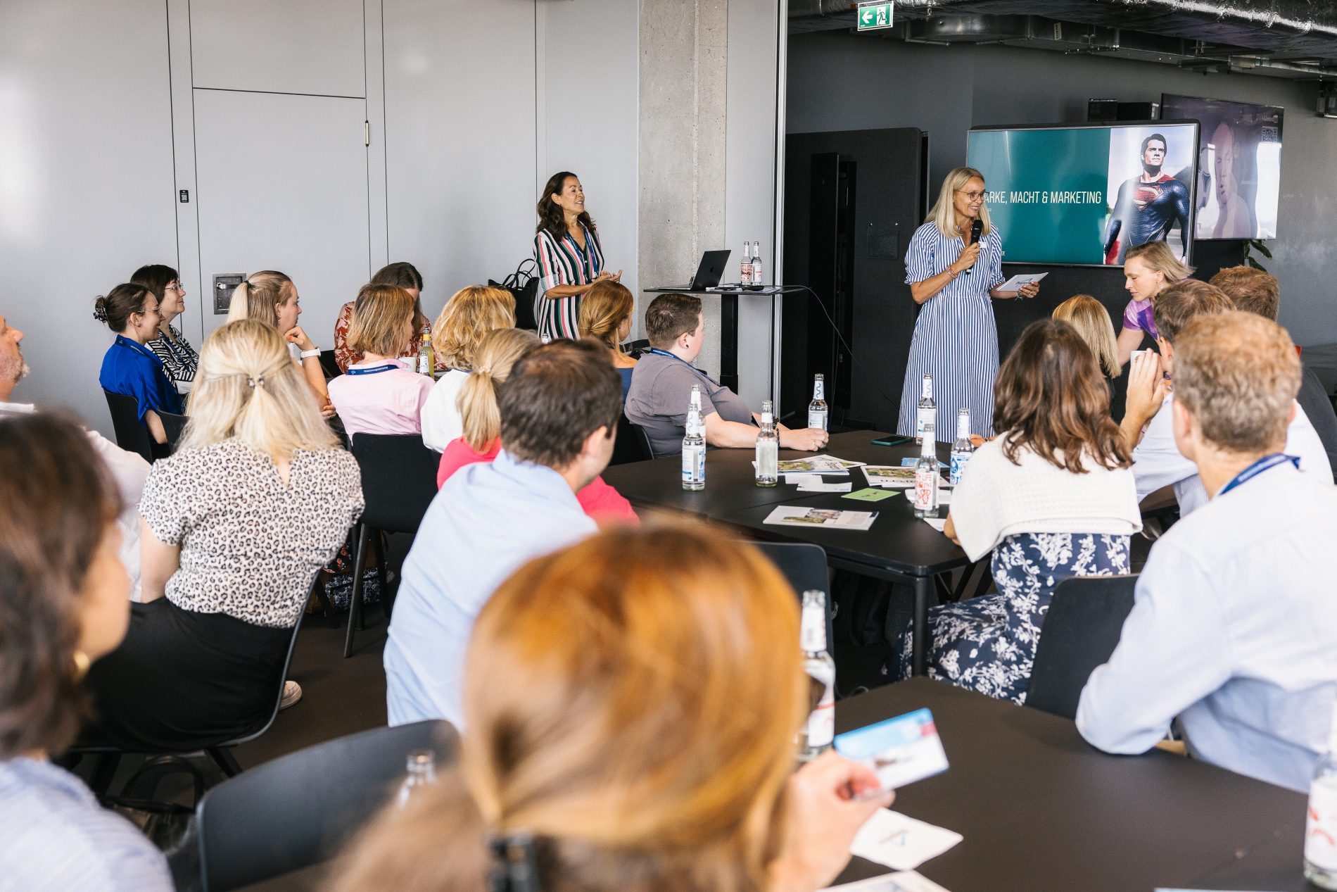 Live Netzwerktreffen 2025: Moderatorin spricht vor Publikum um Tische, Teilnehmer hören zu; Bildschirm zeigt Marketing-Thema im Hintergrund.