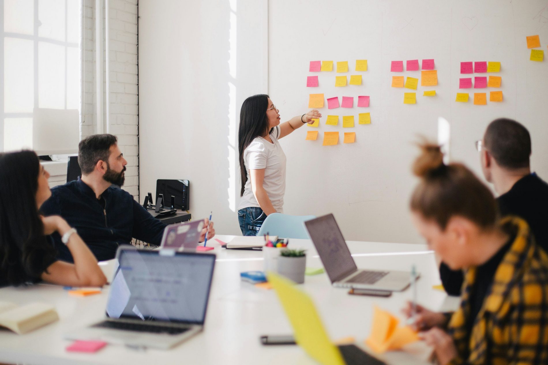 Meeting Mitarbeiter Frau präsentiert Sticky-Notes an Wand; Team mit Laptops diskutiert Arbeitswelt im Tourismus.