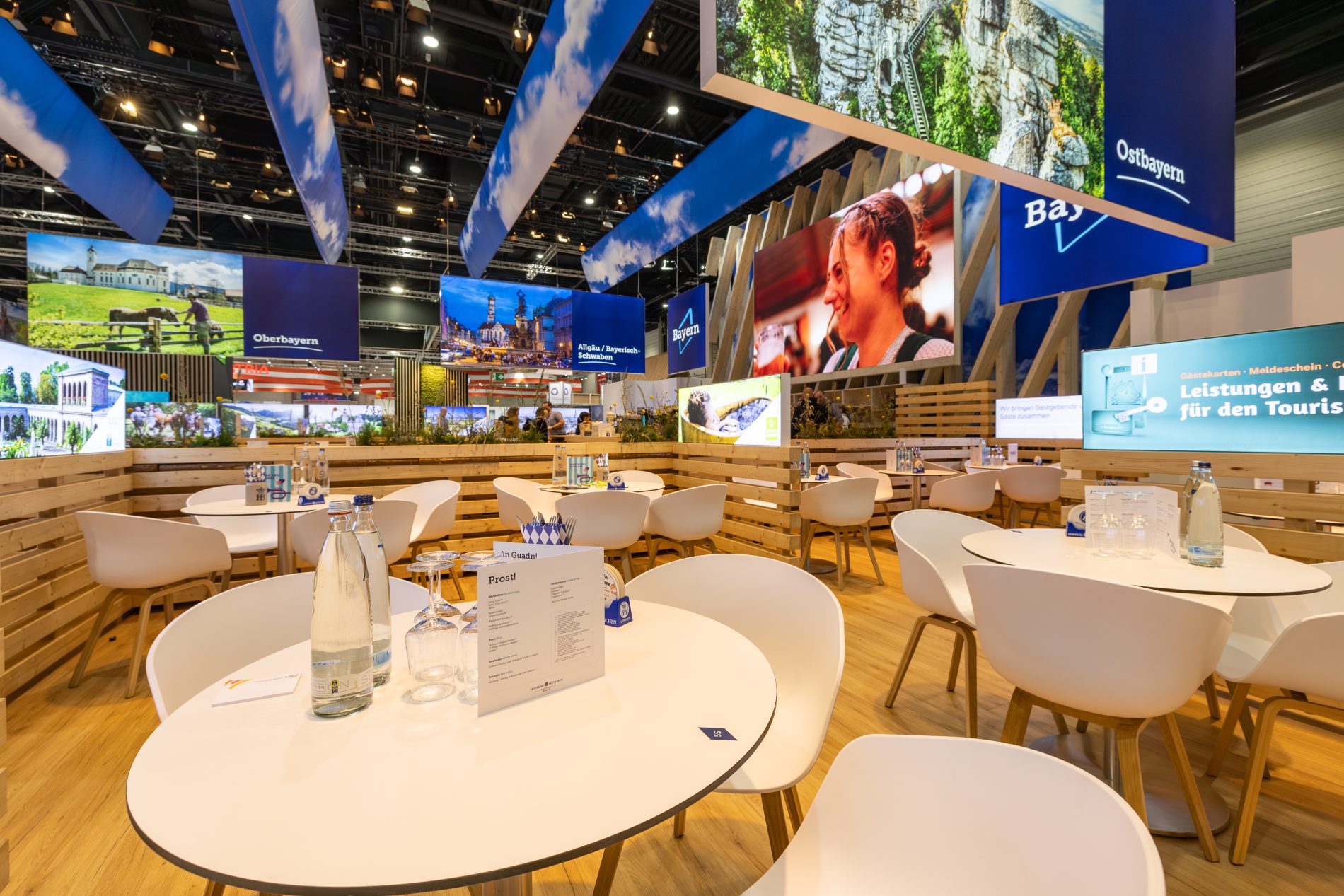 Bayern-Stand auf der ITB Berlin; 4er-Tische zwischen Holzw&auml;nden, Gro&szlig;bildschirm mit Informationen, Banner mit Bayern-Logo.