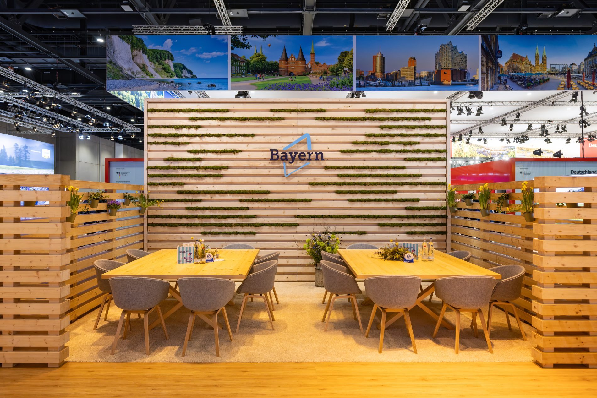 Bayern-Stand auf der ITB Berlin; Lounge mit zwei Tischen und St&uuml;hlen zwischen Holzw&auml;nden mit Pflanzen und Bayern-Logo.