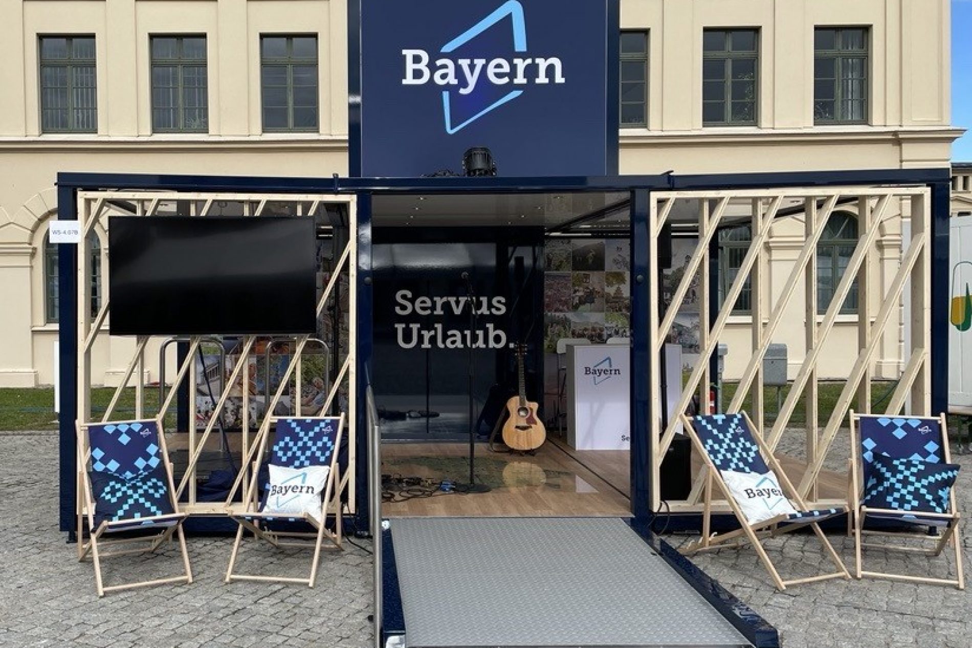 Bayern Live-Event-Cube vor Theatrium Wiesbaden; Eingang mit Rampe, Deckchair-ähnliche Stühle, Gitarre im Innenbereich, Bayern-Logo sichtbar