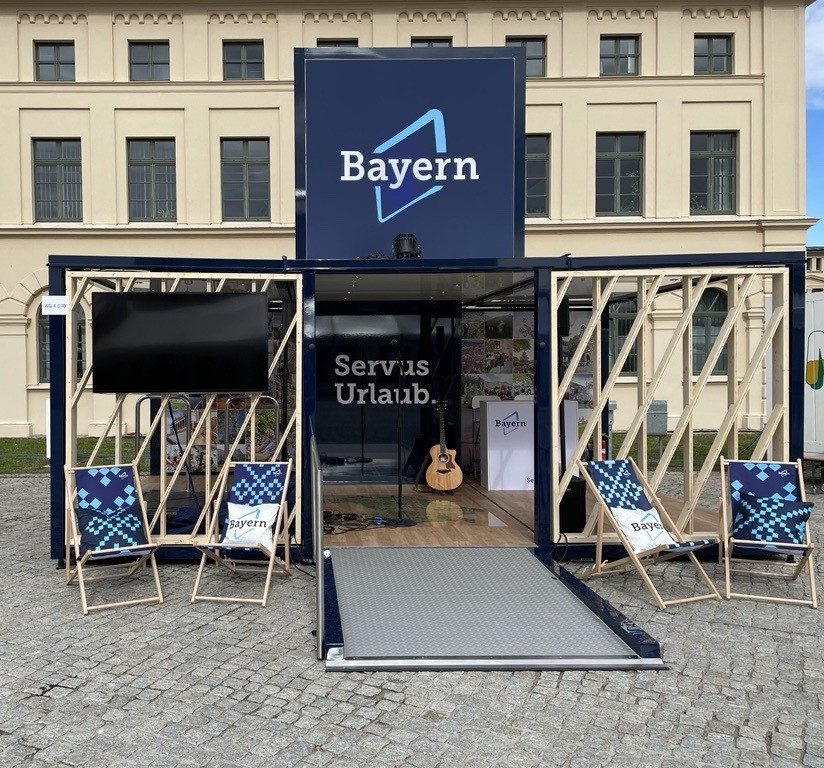 Bayern Live-Event-Cube vor Theatrium Wiesbaden; Eingang mit Rampe, Deckchair-ähnliche Stühle, Gitarre im Innenbereich, Bayern-Logo sichtbar
