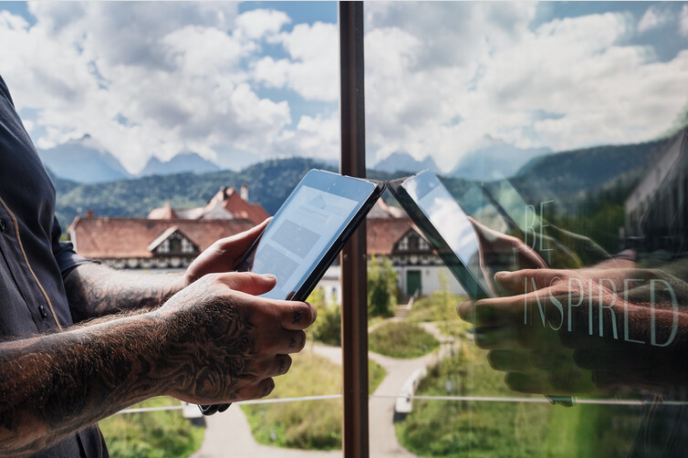 Zwei Hände halten Tablets vor Fenster; Spiegelung einer Berglandschaft mit Häusern, Text Be Inspired sichtbar.