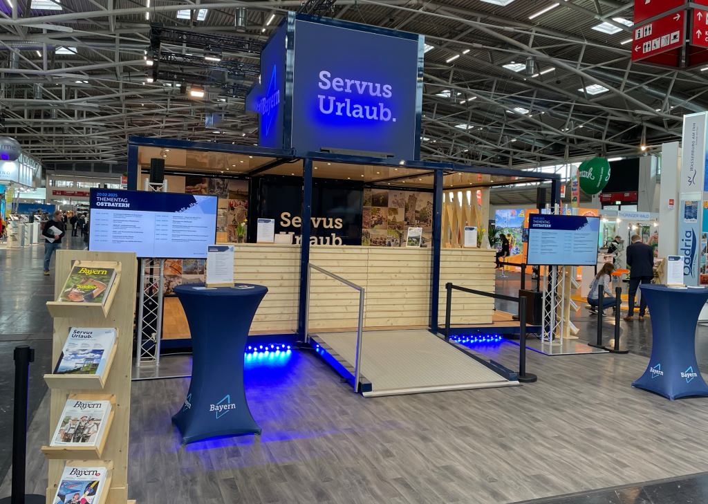 Ausstellerstand mit blau beleuchtetem Servus Urlaub-Logo, Infotafeln, Rampenzugang und Broschürenständer auf Messe.