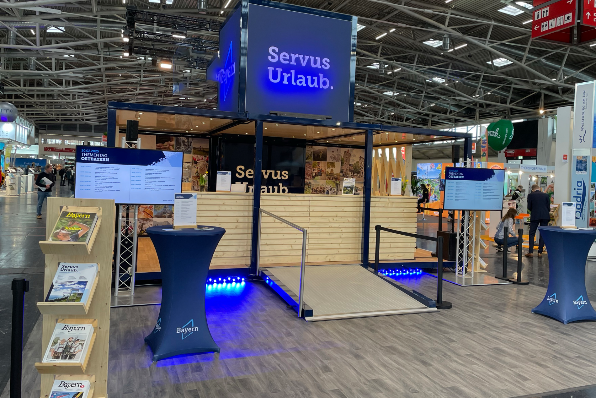 Event-Cube2 Ausstellerstand mit blau beleuchtetem Servus Urlaub-Logo, Infotafeln, Rampenzugang und Broschürenständer auf Messe.