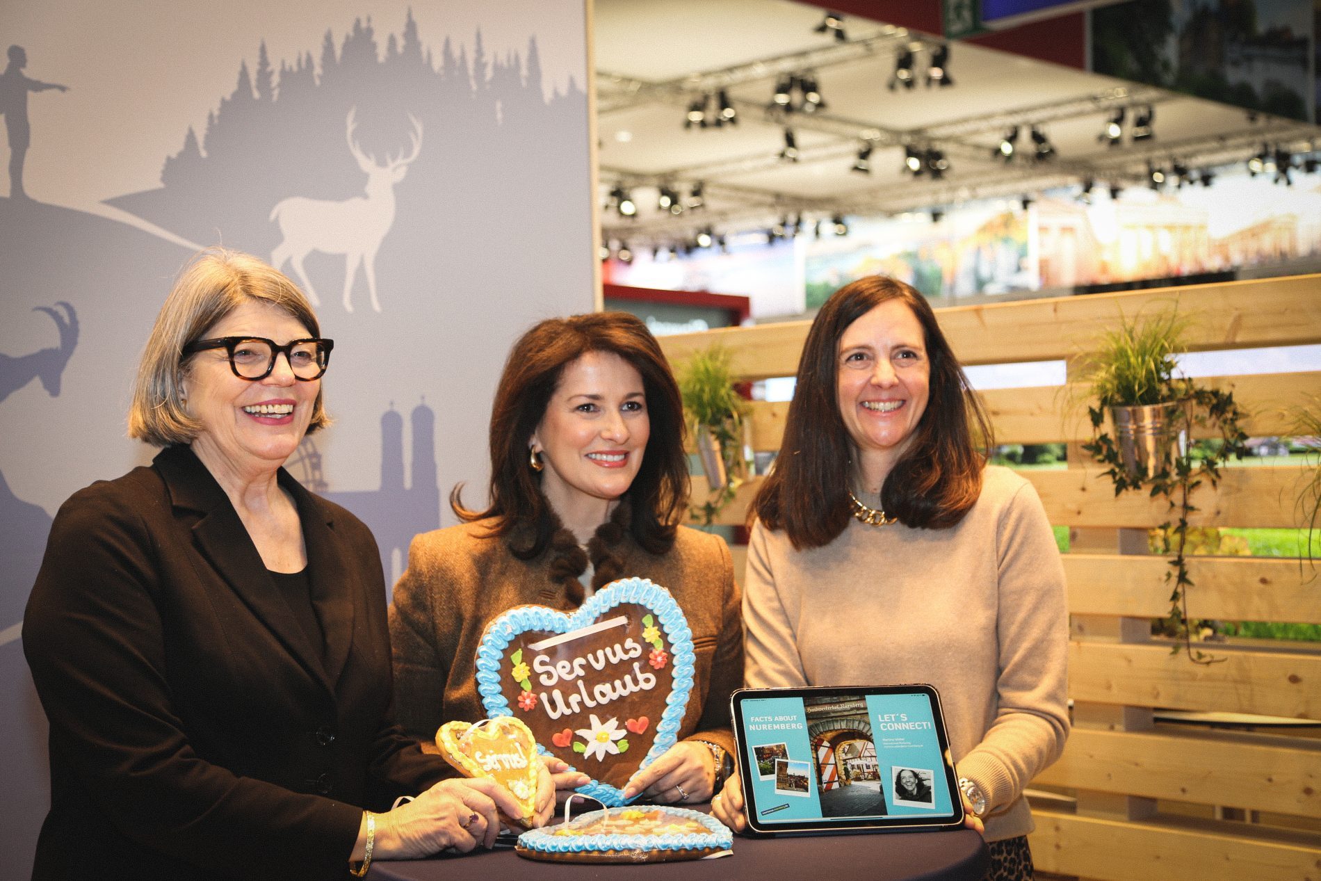 Drei Frauen am Bayern-Stand der ITB Berlin 2025 halten ein Zuckerkuchen-Herz und ein Tablet.