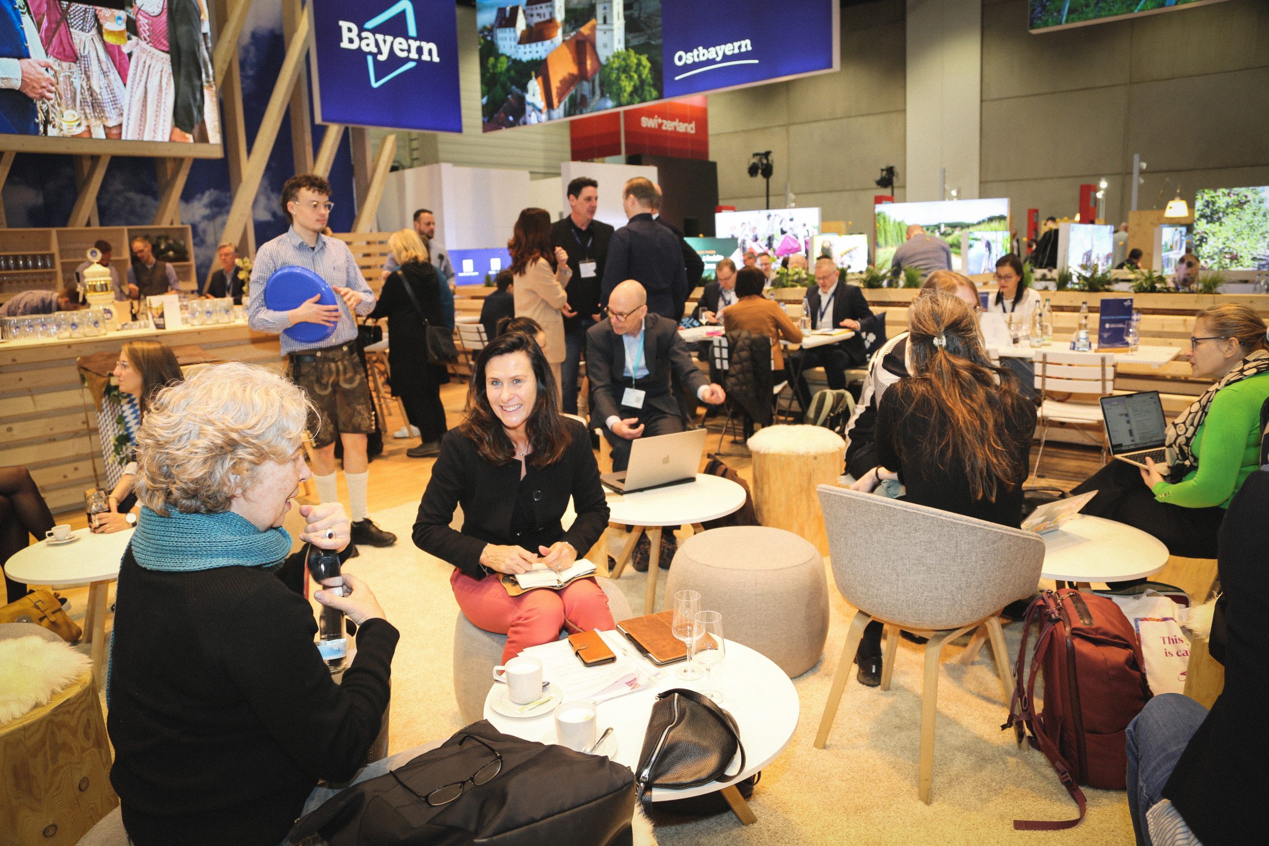 Bayern-Stand auf der ITB Berlin 2026; Besucher sitzen, sprechen, arbeiten an Laptops, Getr&auml;nke und Tische im Lounge-Bereich.
