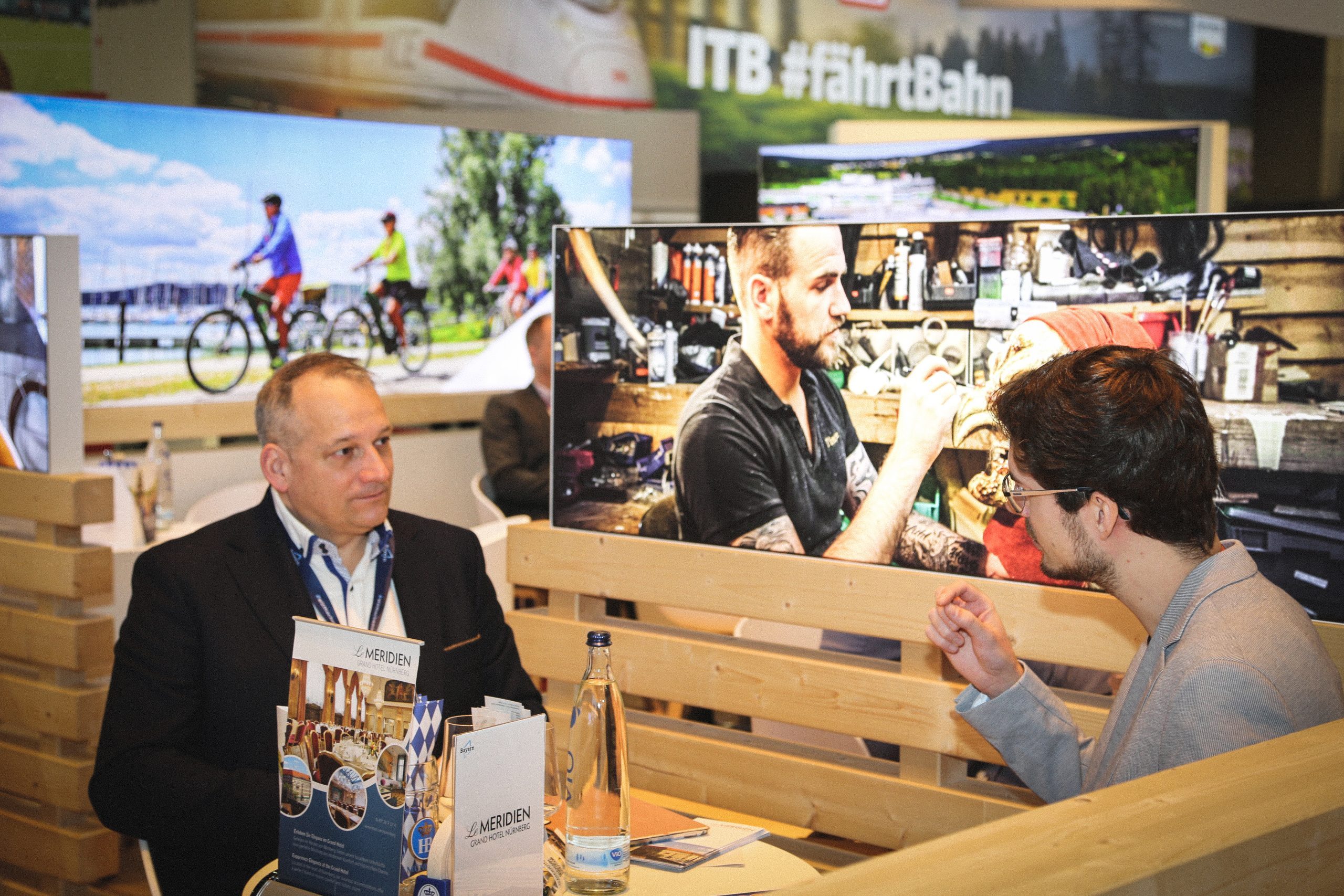 Zwei M&auml;nner im Gespr&auml;ch am Bayern-Stand auf der ITB Berlin 2025; Infomaterial, Wasserflasche, Holzwand, Hintergrundfotos