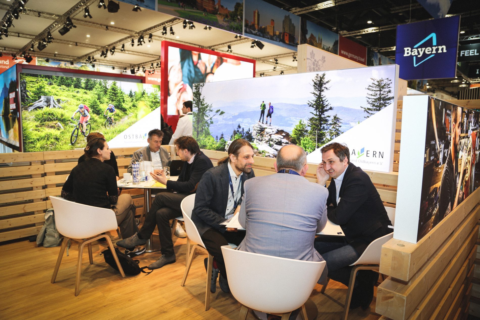 Mehrere Geschäftsleute sitzen am Bayern-Stand der ITB Berlin 2025; Holzumrandung, Bergpanorama im Hintergrund.