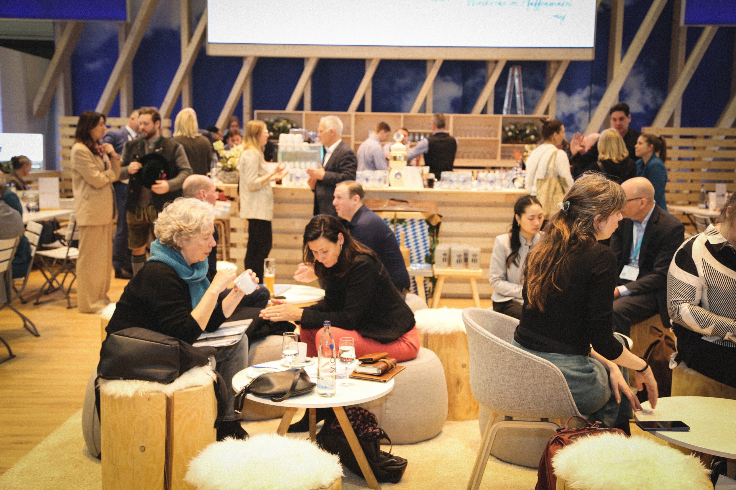 Menschen bei einer Veranstaltung an einer Holzwand-Bar, stehen und sitzen im Gespr&auml;ch, Getr&auml;nke und Snacks sichtbar.