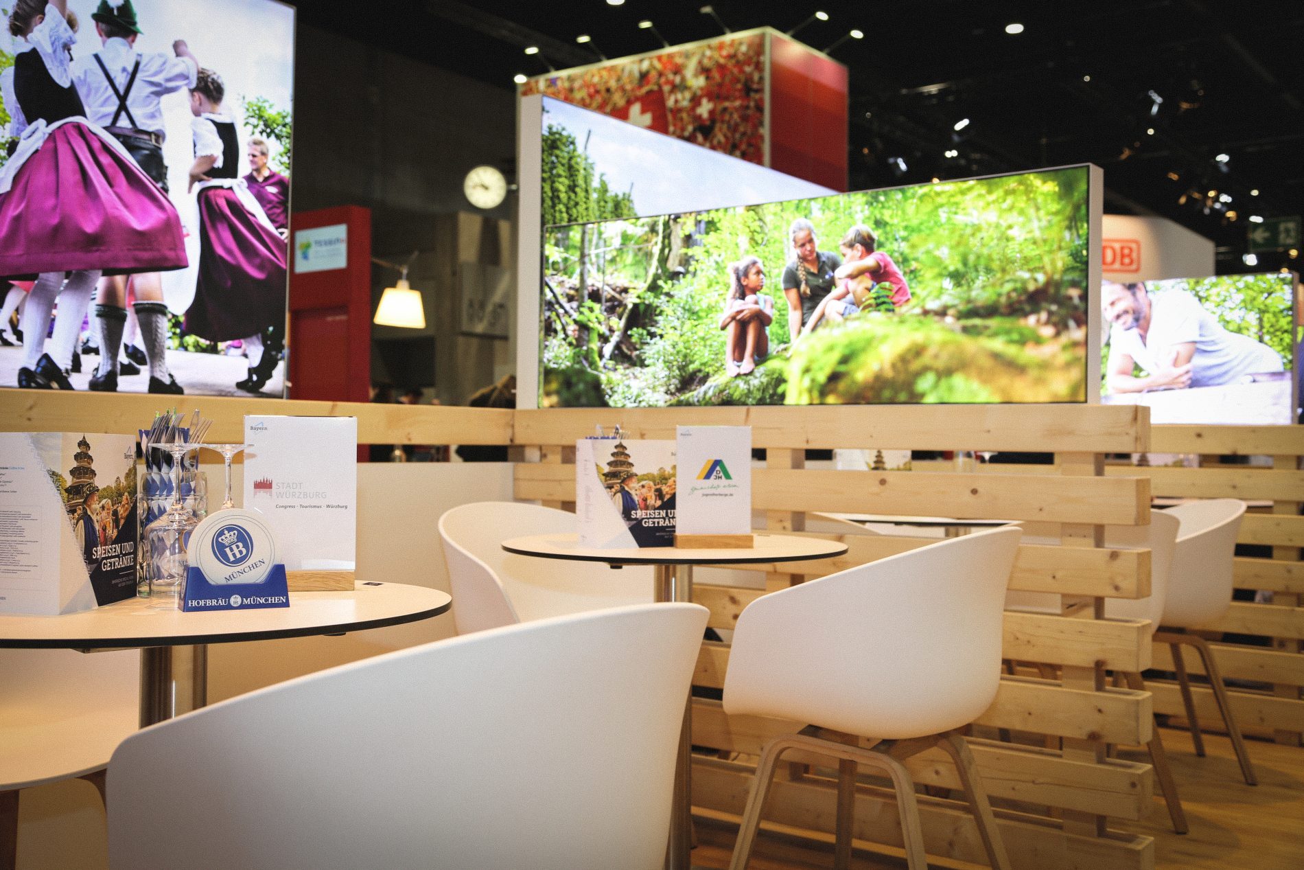 ITB-Modul 4 mit 2er Tisch: runde Tische, weiße Stühle, Holzwand-Trennwand, Informationalsmaterial, große Bildschirme im Hintergrund.