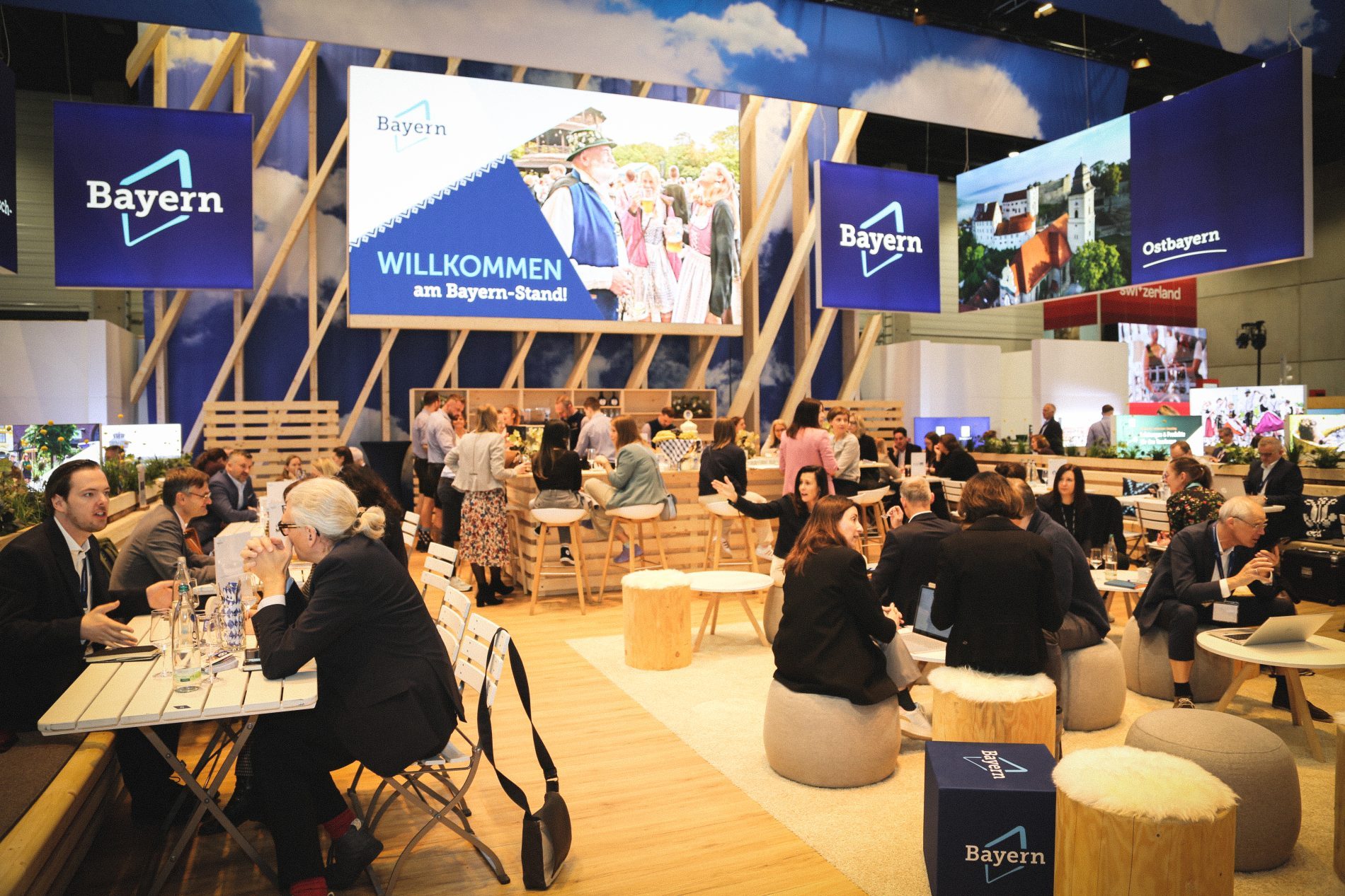 Bayern-Stand auf der ITB Berlin 2026; Besucher sitzen an Holztischen, Banner WILLKOMMEN am Bayern-Stand und Bayern-Logos.