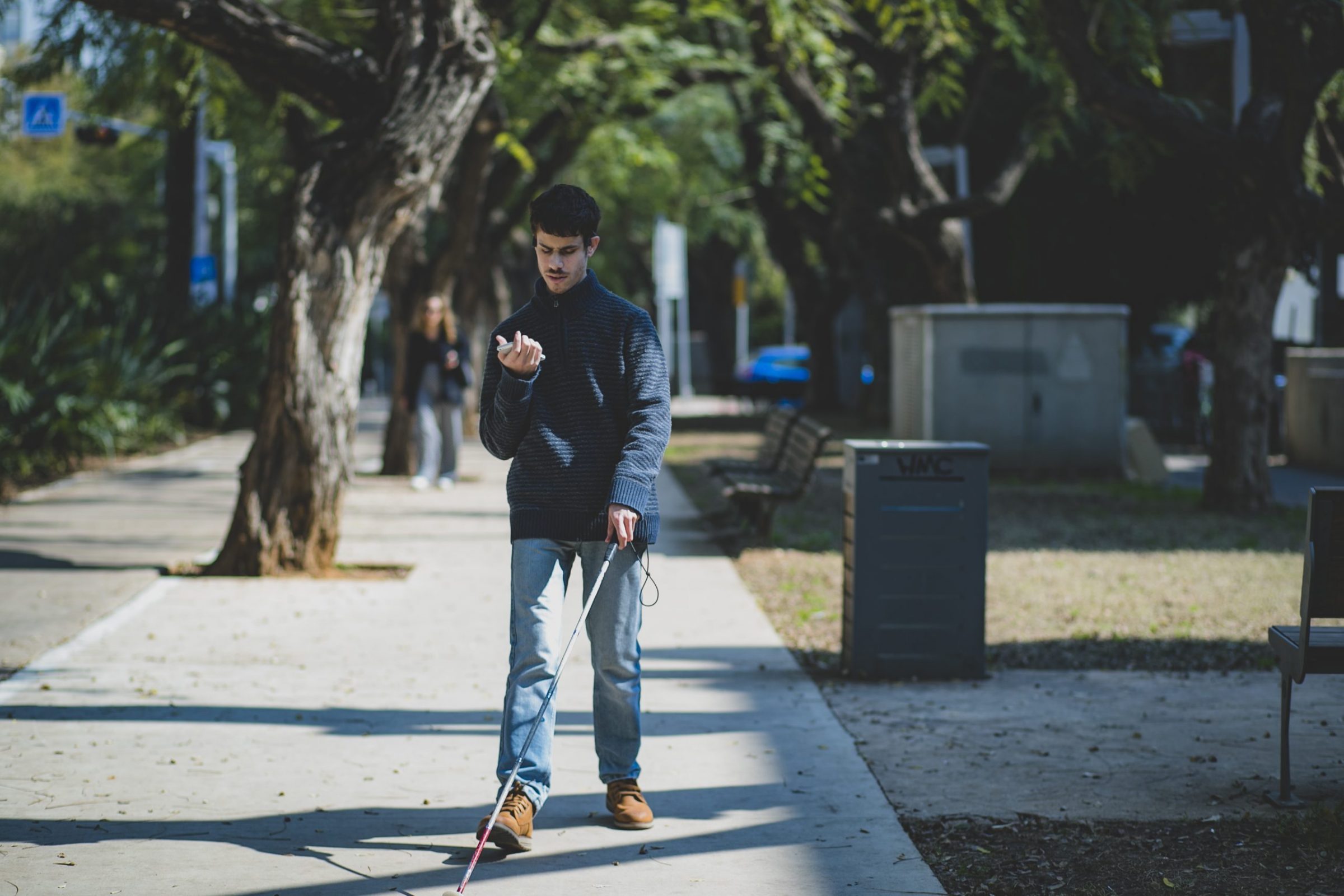 RightHear App Ein junger Mann mit Blindenstock geht auf einem Gehweg im Park entlang. Er trägt einen dunklen Pullover und helle Jeans, hält ein Smartphone in der Hand und nutzt den Stock zur Orientierung. Im Hintergrund sind Bäume, Bänke und eine weitere Person erkennbar.