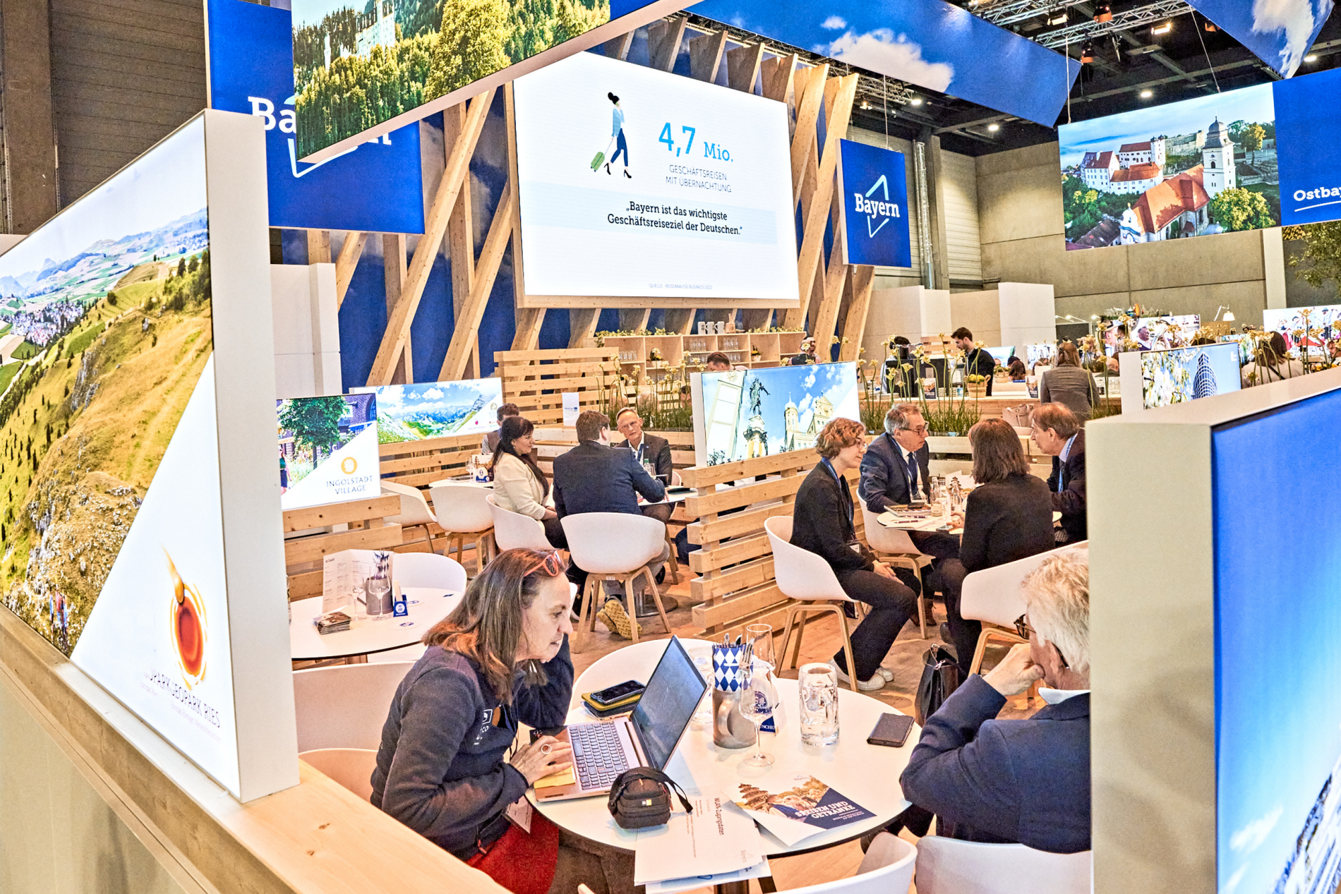 Bayern-Stand auf der ITB Berlin; Gruppen an 4er-Tischen hinter Holzwand, Großbildschirm mit Informationen, Banner mit Bayern-Logo.