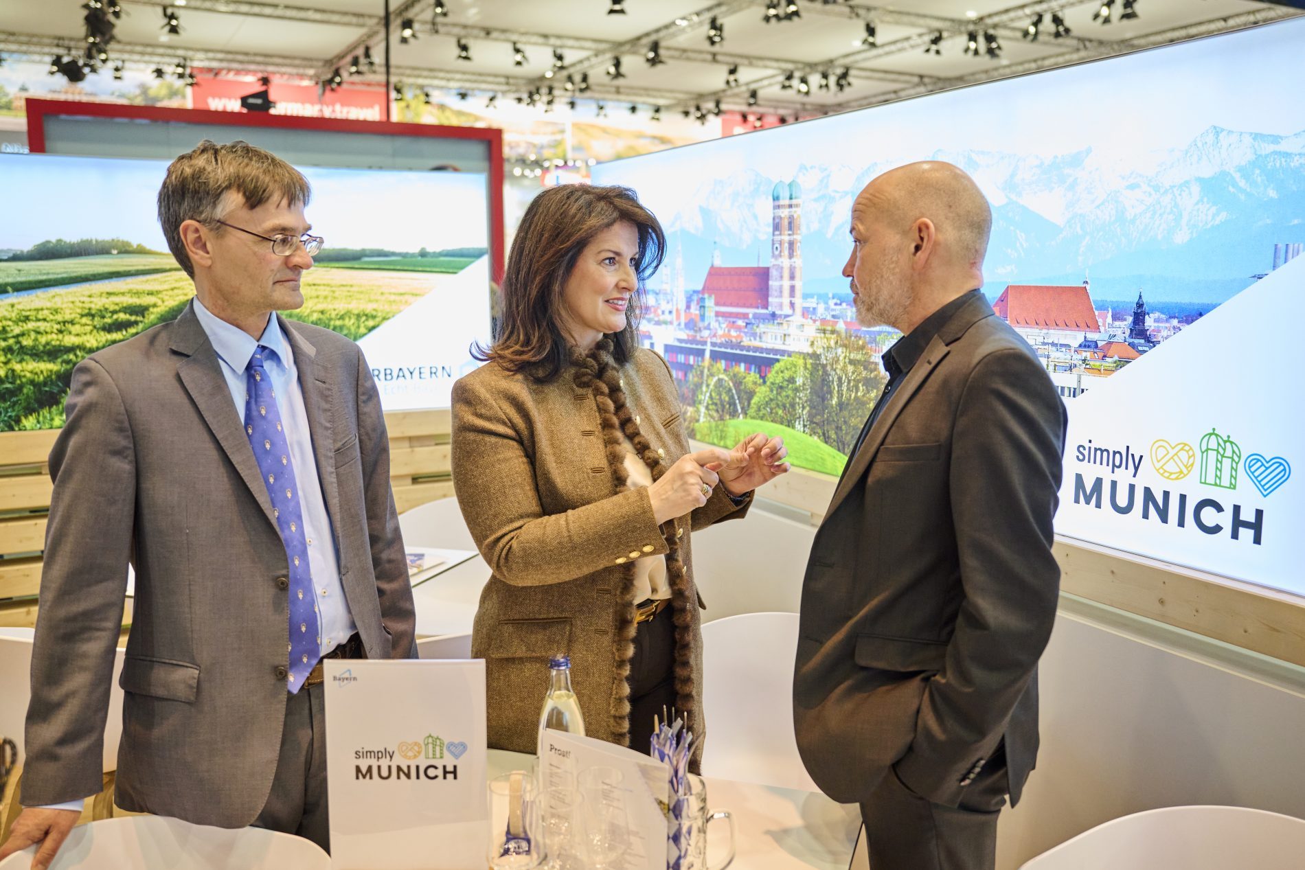Drei Personen im Bayern-Stand auf der ITB Berlin 2025/2026 im Gespräch; Hintergrund Münchner Stadtpanorama mit Alpen