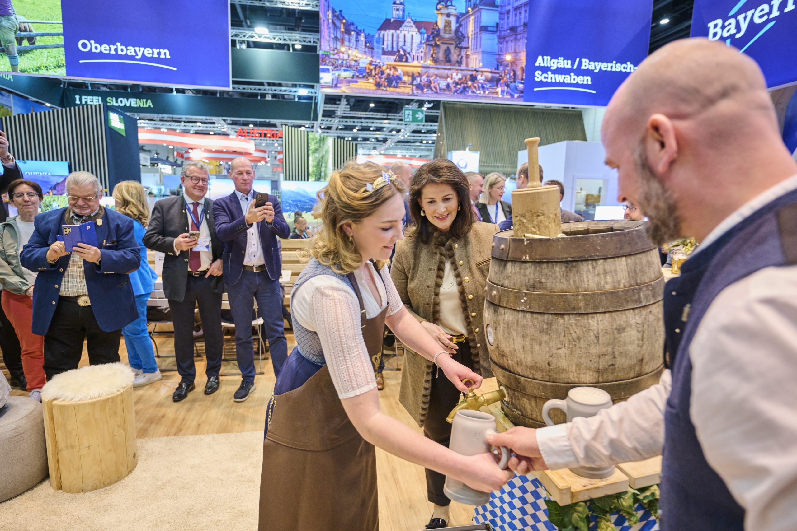 Bayern-Stand auf der ITB Berlin 2025; Frau f&uuml;llt aus einem Holzfass Krug, weitere Besucher beobachten.