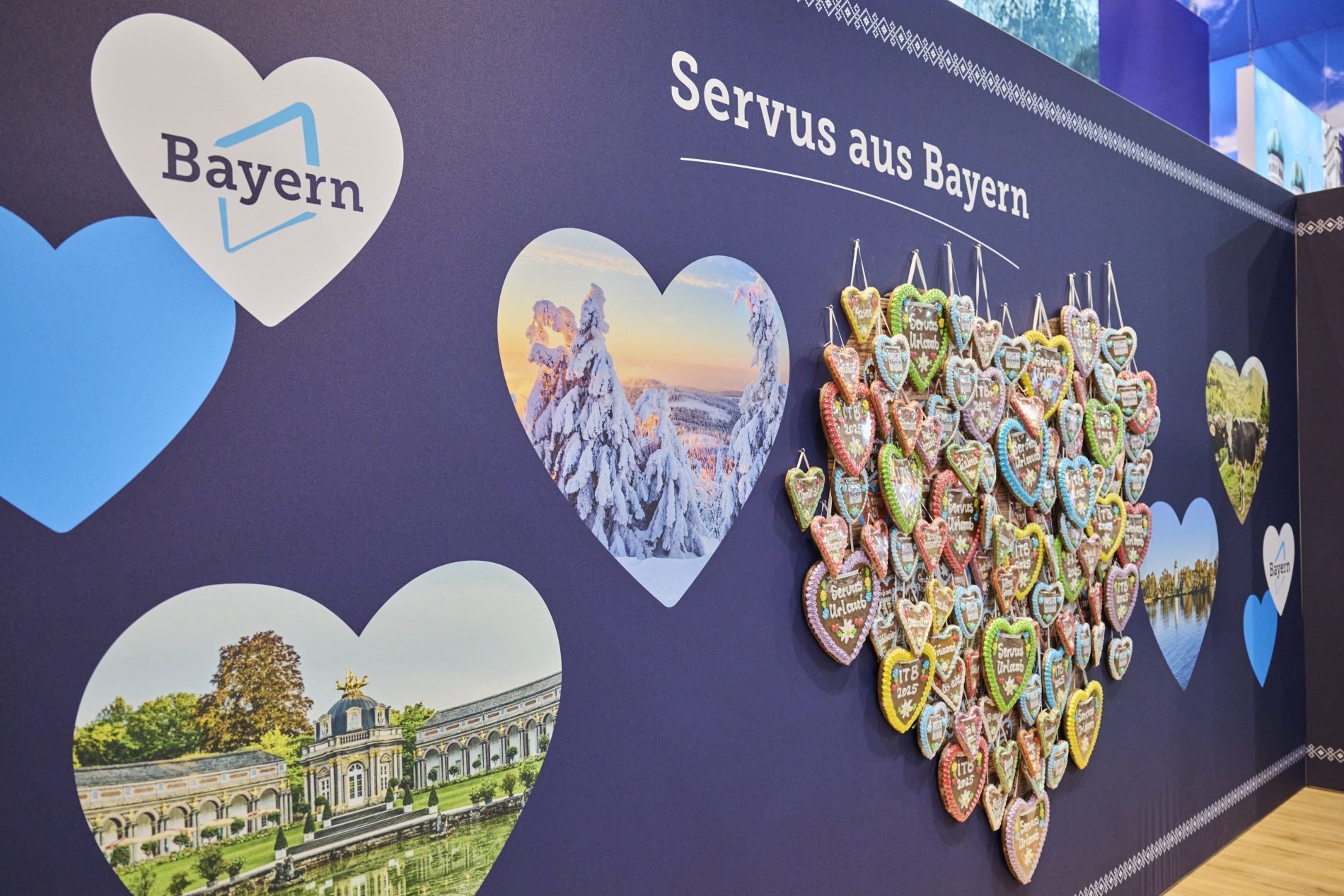 Bayern-Stand ITB Berlin 2025: Wand mit Herzen, Landschaftsbildern und Lebkuchenherzen; Text Servus aus Bayern.