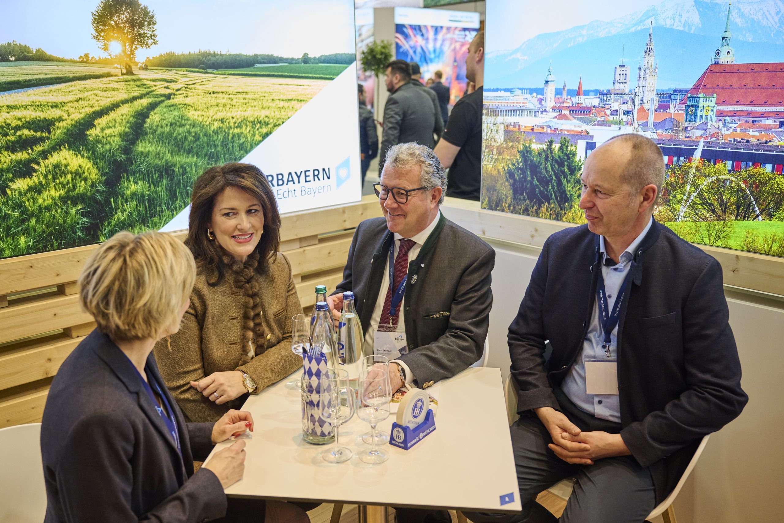 Vier Personen im Gespr&auml;ch am Bayern-Stand auf der ITB Berlin 2025; Getr&auml;nke auf dem Tisch, Hintergrund mit Landschaftsbildern.