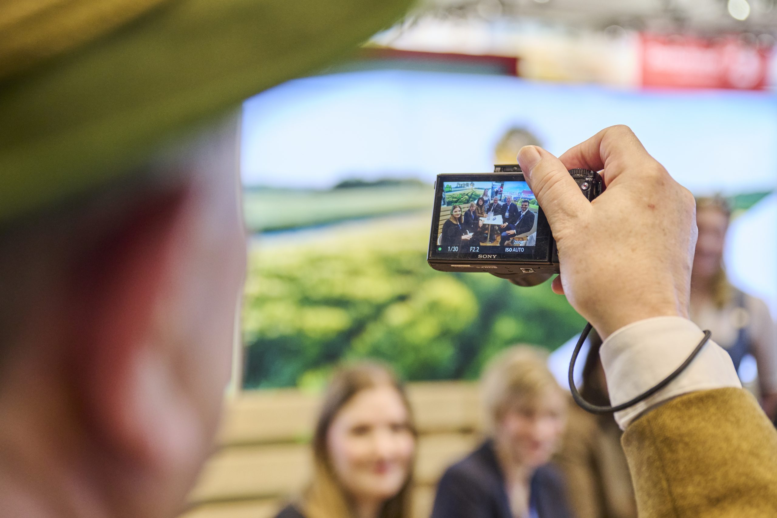 Hand h&auml;lt kompakte Kamera; Display zeigt Gruppe von Menschen vor Bayern-Stand bei der ITB Berlin 2025