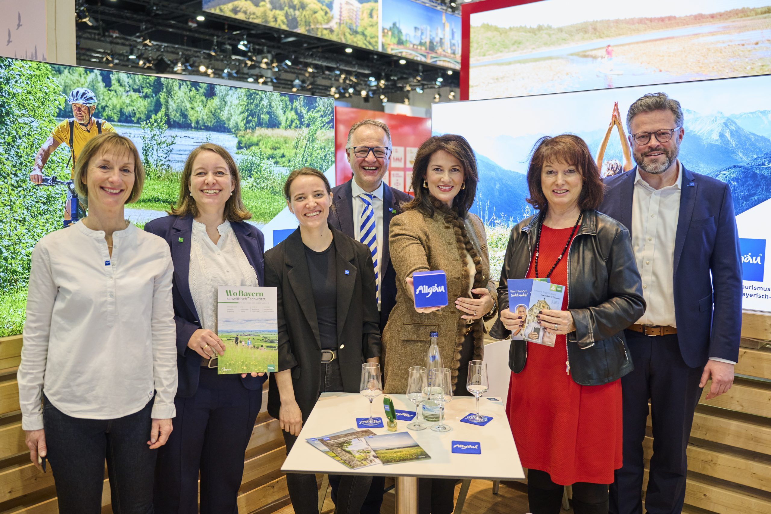 Gruppe von sieben Personen am Bayern-Stand ITB Berlin 2025, halten Brosch&uuml;ren vor Hintergrundfotos mit Naturlandschaften.
