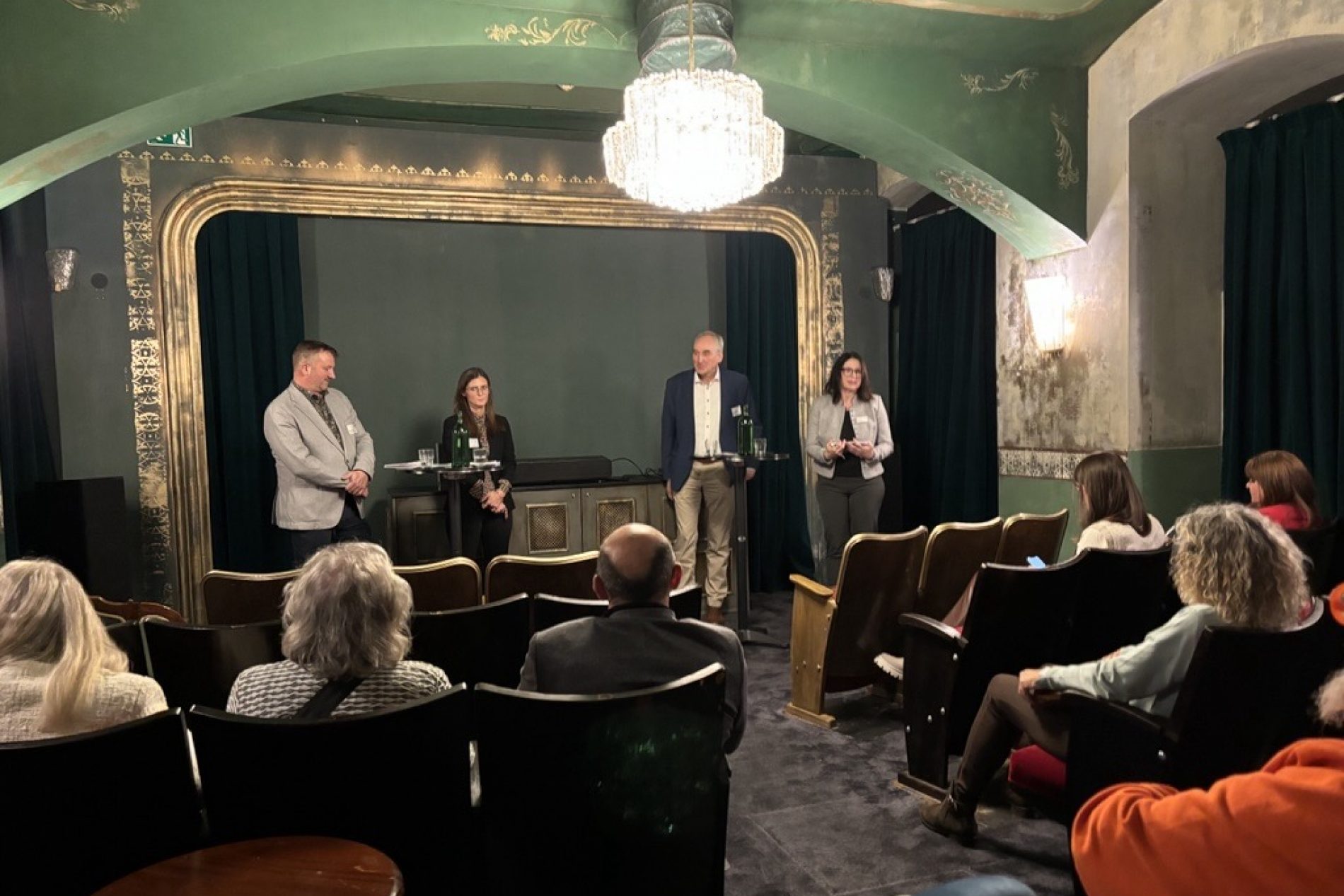 Vier Personen stehen auf einer kleinen B&uuml;hne im gr&uuml;nen Saal mit Kronleuchter; Publikum sitzt in Sesseln und verfolgt eine Podiumsdiskussion.