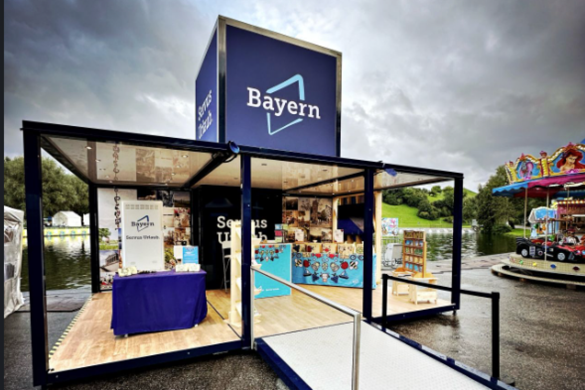 Ausstellerstand Bayern auf einem offenen Event-Cube bei OutdoorWelt K&ouml;ln; Rampe, Glasbox, blaue Beschilderung, Infotisch, Karussell im Hintergrund.