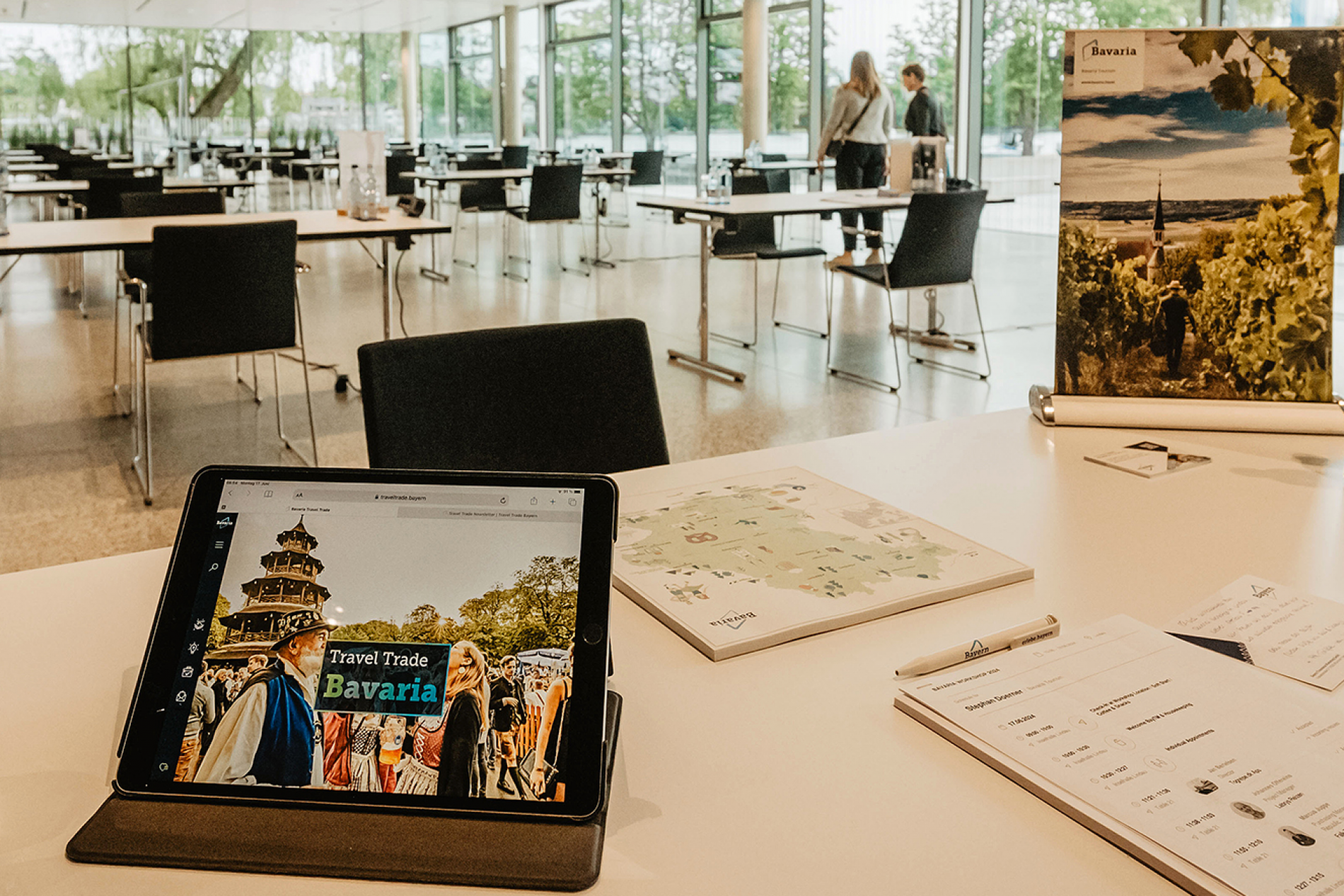 Tablet zeigt Travel Trade Bavaria auf einem Tisch; Hintergrund: heller Konferenzraum mit St&uuml;hlen und einem Bayern-Poster.