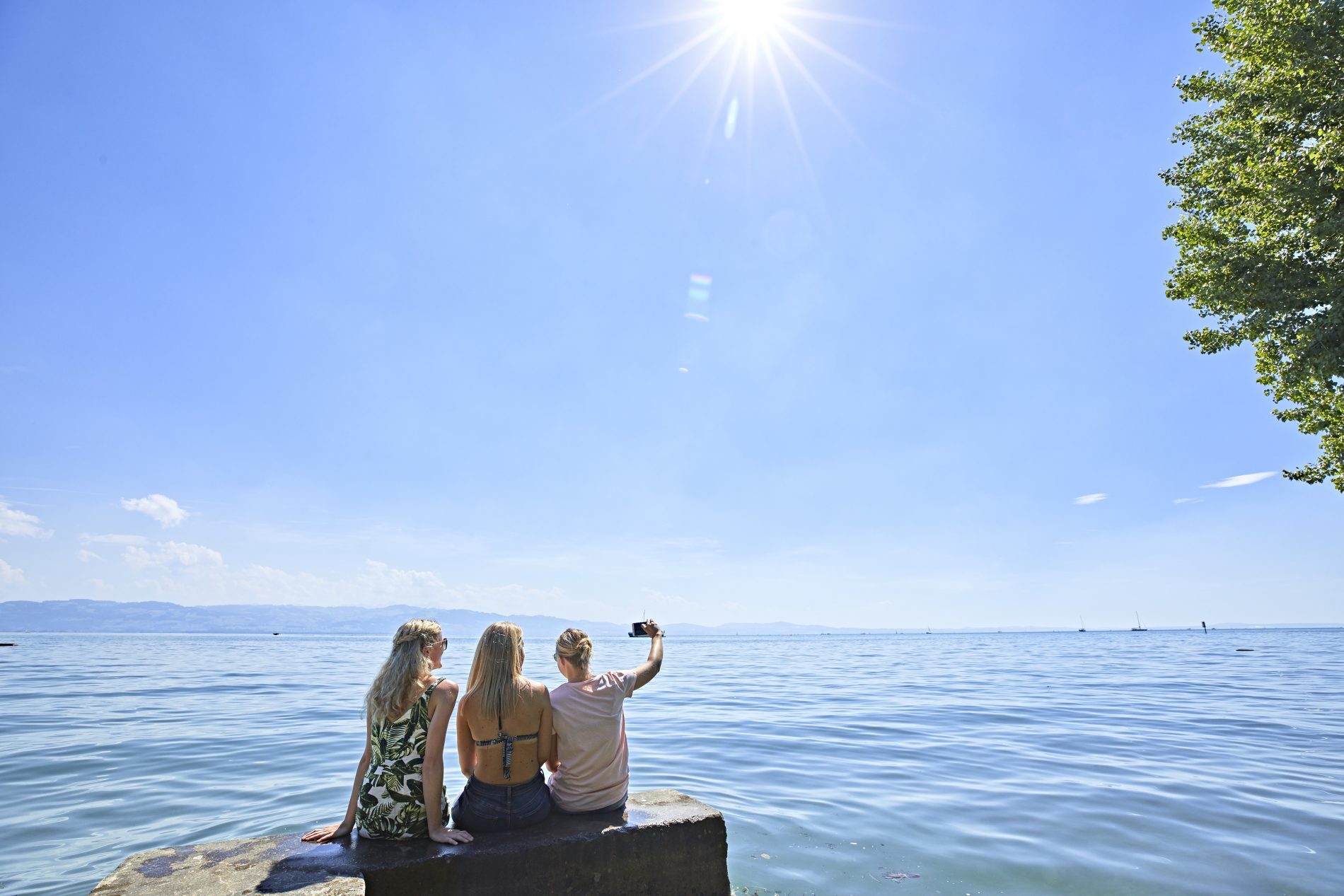 Drei Frauen sitzen auf einer Steinplatte am Bodensee in Nonnenhorn, machen Selfie, klarer Himmel und ruhiges Wasser.