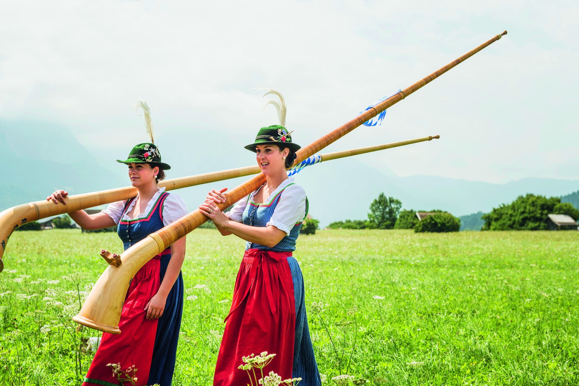 Zwei Frauen in Dirndl tragen lange Alphörner durch eine grüne Wiese vor Bergen; Sponsored Teaser im Netzwerk-Update