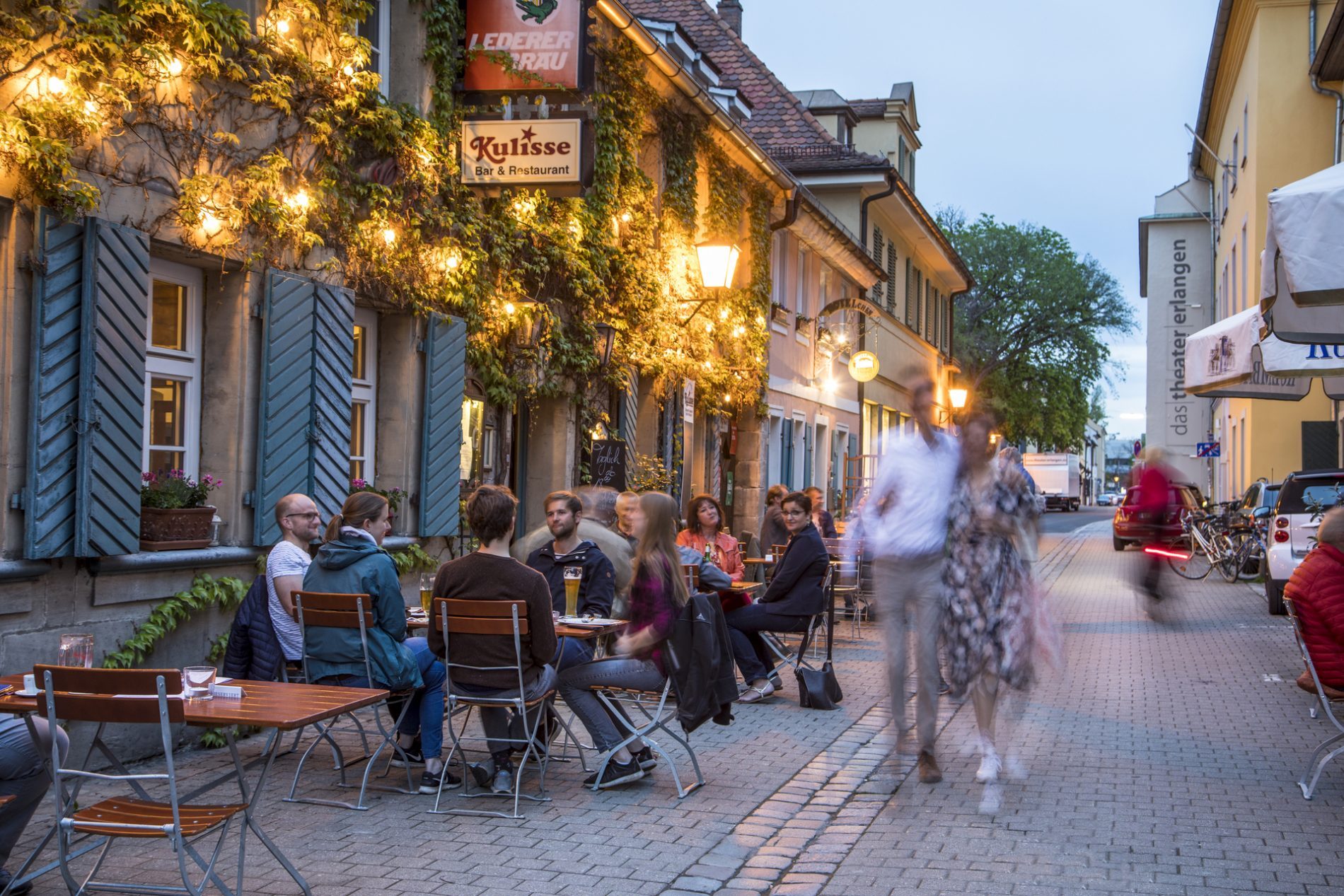 Abendliches Stra&szlig;encaf&eacute; in Erlangen mit G&auml;sten an Tischen, Lichterketten an der Fassade und Schild Kulisse Bar & Restaurant.